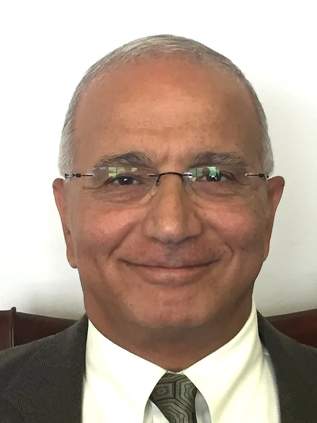 Dr. Joseph Danial, with short gray hair and glasses, wearing a dark suit, white shirt, and patterned tie, smiling in front of a plain white wall.