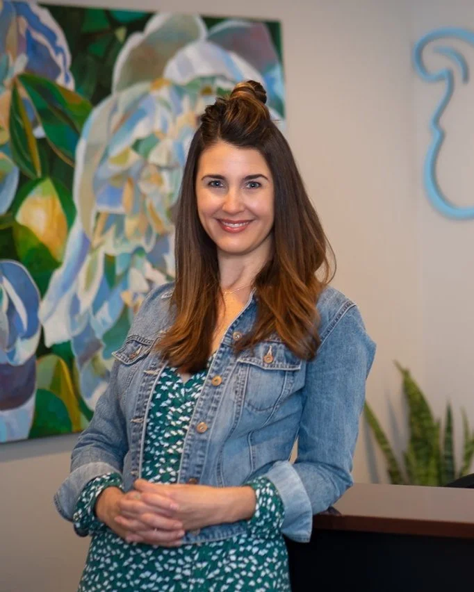 Kellie Wicklund, with long brown hair, styled in a half-up bun, smiling while standing indoors in front of abstract art and a plant, wearing a denim jacket over a green patterned dress.