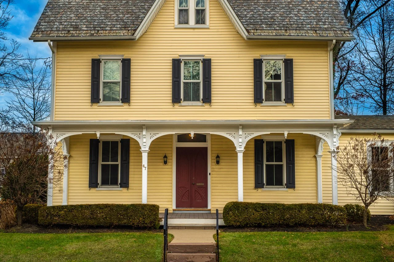 67 Byberry Rd, Yellow two-story house with black shutters, a red front door, and a porch supported by white columns, bordered by bushes and a grassy lawn.