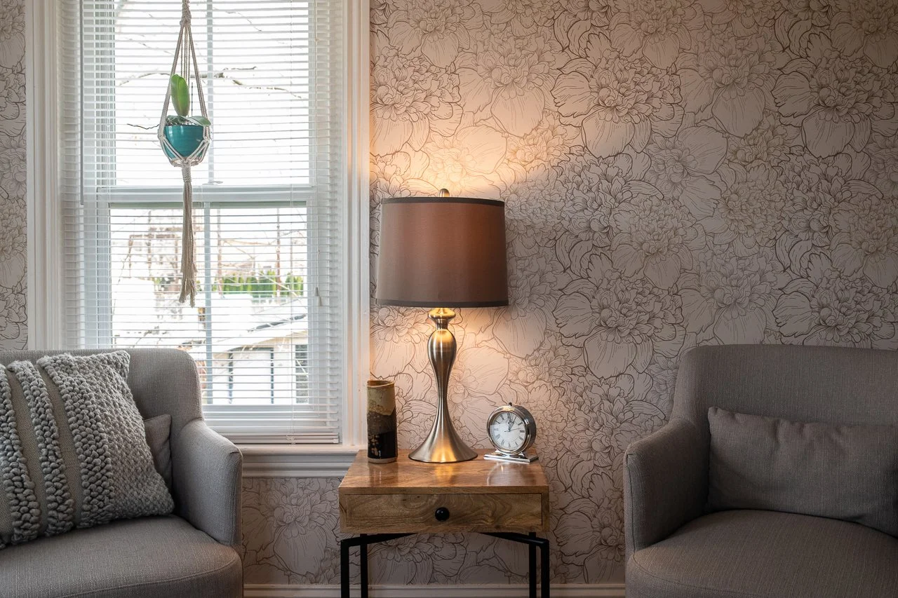 67 Byberry Rd, Living room with floral wallpaper, wooden side table with a lamp, clock, and decorative item, two gray armchairs, one with a textured pillow, window with blinds, and a hanging plant outside.
