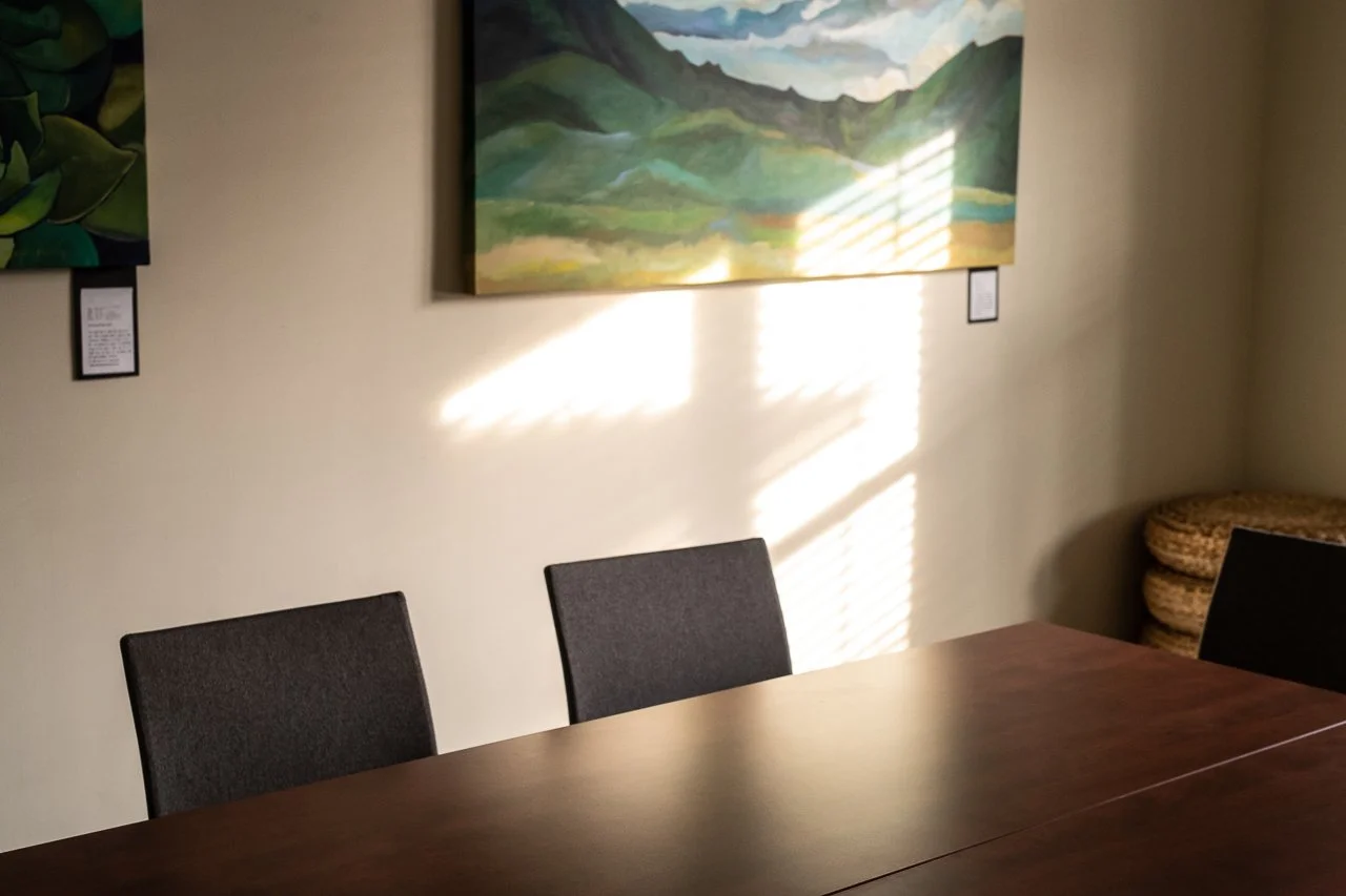 A conference room with a wooden table, three gray chairs, a landscape painting on the beige wall, and sunlight streaming through window blinds, creating light patterns on the wall.