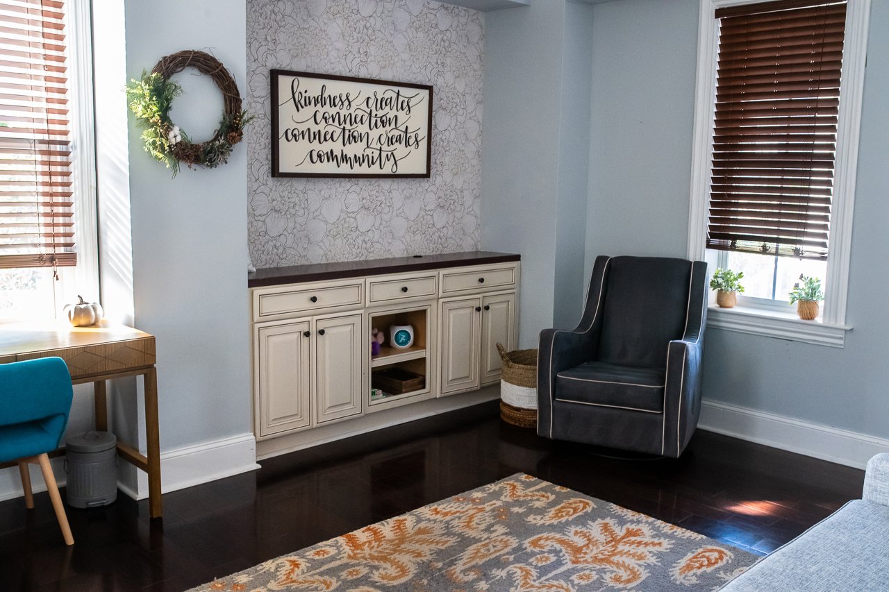 67 Byberry Rd, A cozy living room with a black armchair, dark hardwood flooring, light blue walls, a white cabinet with drawers, a floral wallpaper accent wall, and decorative items including a wreath, potted plants, and a framed quote.