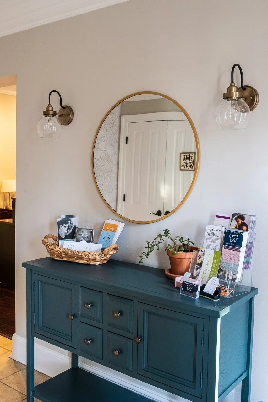 A blue sideboard with a mirror above it, two wall-mounted light fixtures on each side, and informational brochures and a potted plant on top.