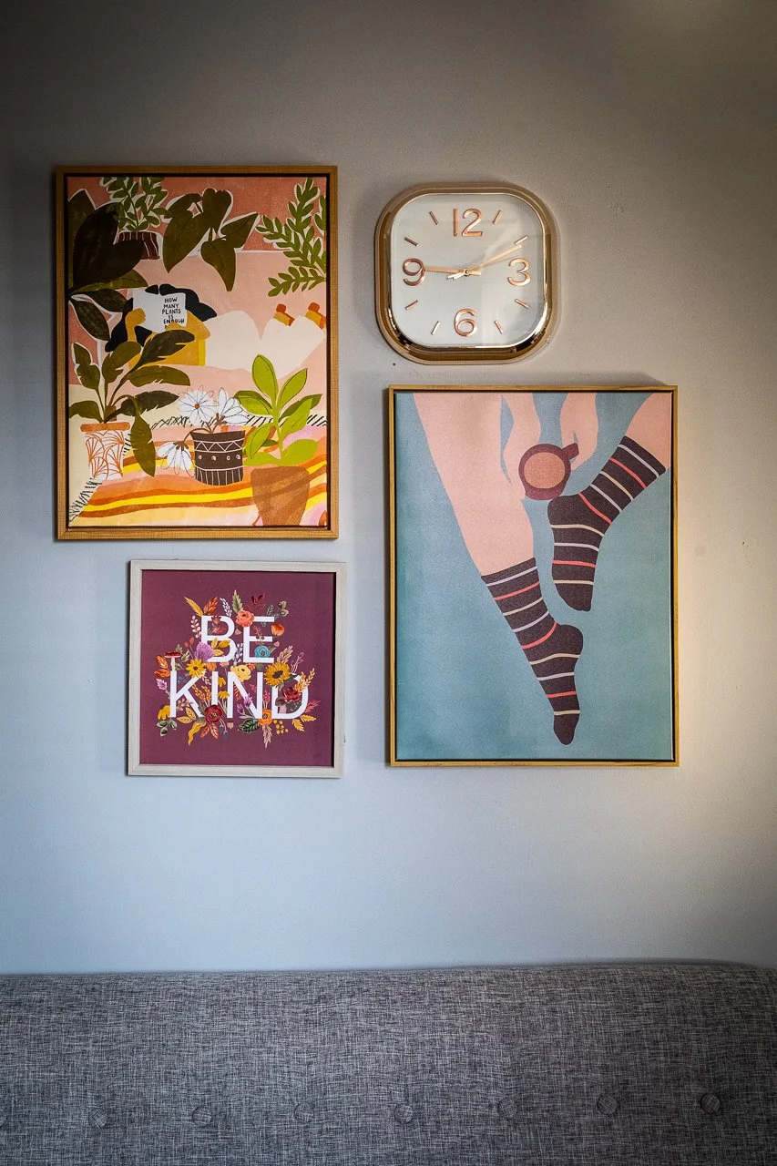 A wall decorated with four framed art pieces and a clock. The art includes a plant scene, a 'BE KIND' floral design, and a drawing of legs wearing striped socks and sneakers. The clock shows the time as approximately 2:12.
