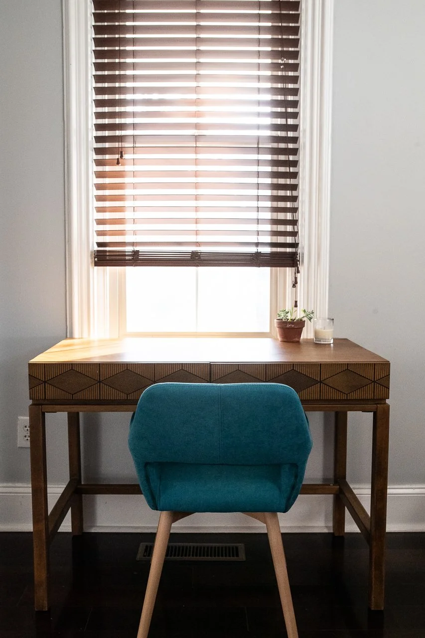 67 Byberry Rd, A wooden desk with geometric carvings on the front, a teal upholstered chair, a small potted plant, and a candle on the desk, in front of a window with brown horizontal blinds partially open letting light in.