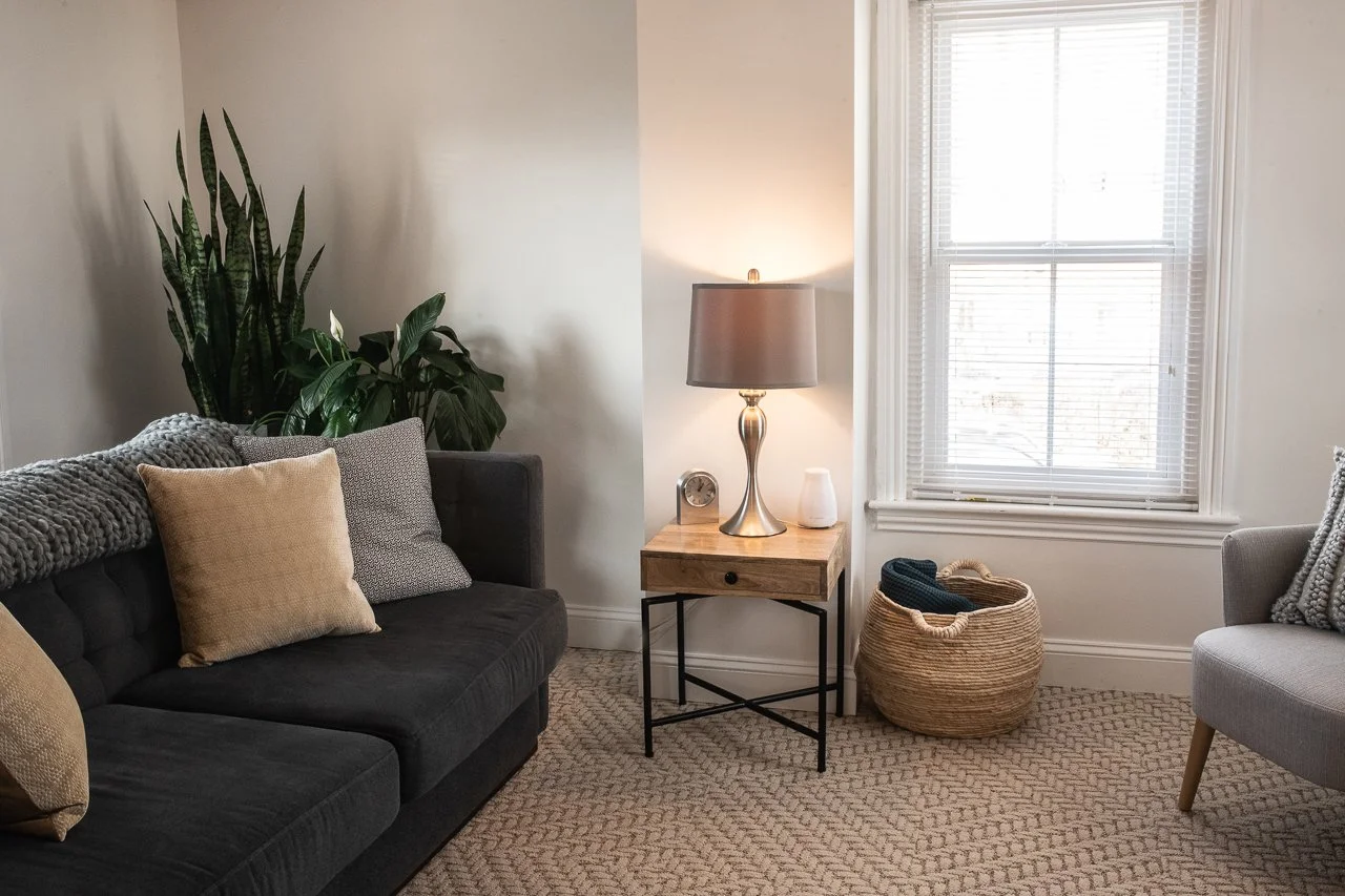 67 Byberry Rd, Living room with a dark gray sofa, beige and gray pillows, a large green plant, a wooden side table with a lamp, a small clock, and a white diffuser, a wicker basket with blankets, and a gray armchair by a window with blinds.
