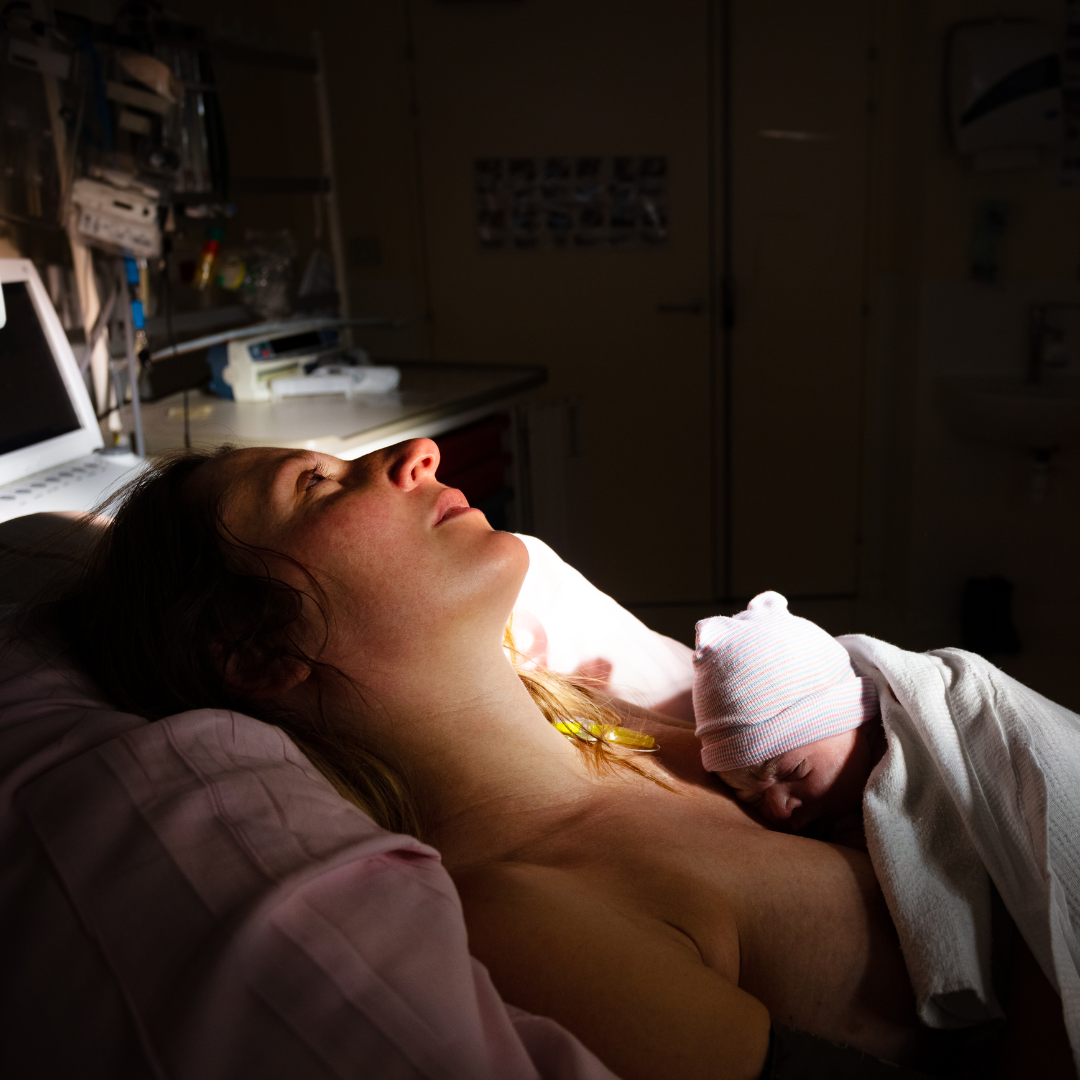 Mother postpartum holding baby with pink and blue striped hat.  Mother is gazing at the ceiling with blank stare.