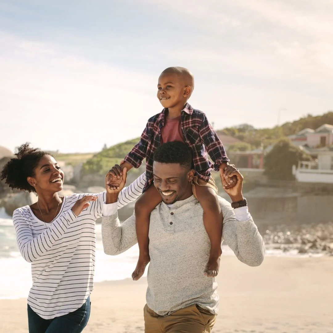 A family walks on the beach on a sunny day. There is a child riding on the black man's shoulders and a black female is supporting the child's back. Everyone is smiling.