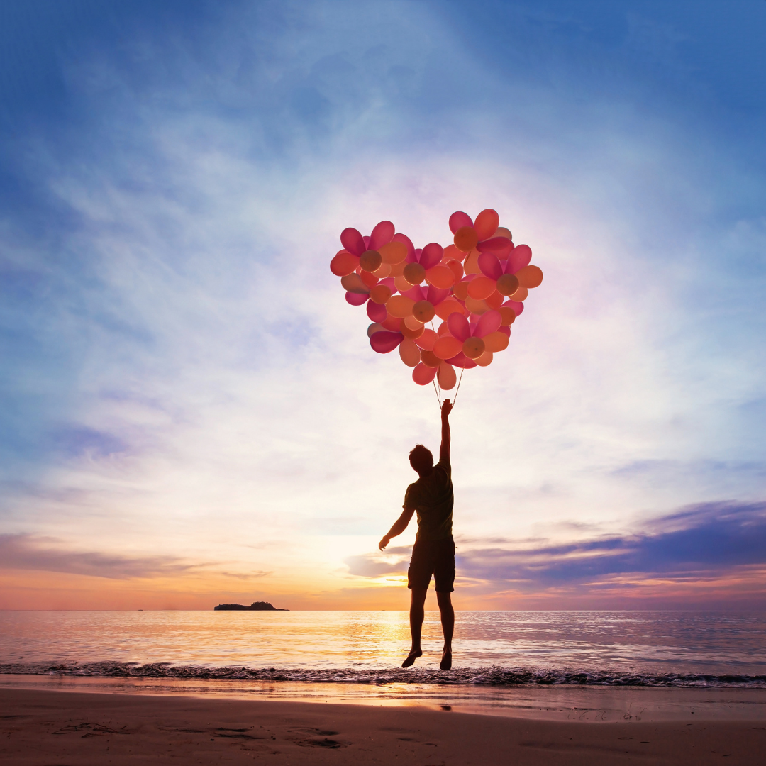 Image of adult holding onto a bunch of balloons that are shaped like a heart while rising up in the air.  The background is a sunrise.