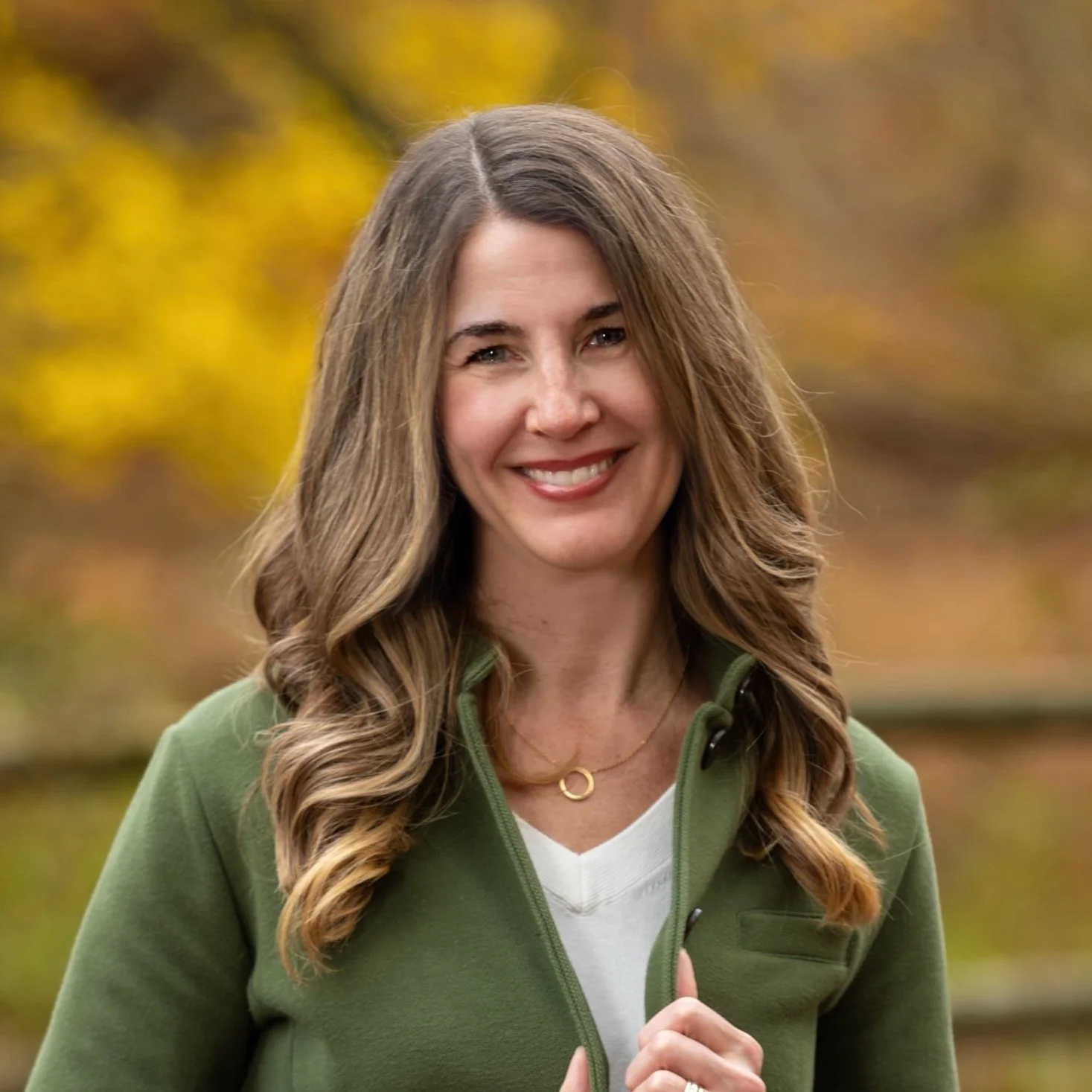 Kellie Wicklund, with long, wavy brown hair smiling outdoors, wearing a green jacket over a white shirt, with autumn foliage in the background.
