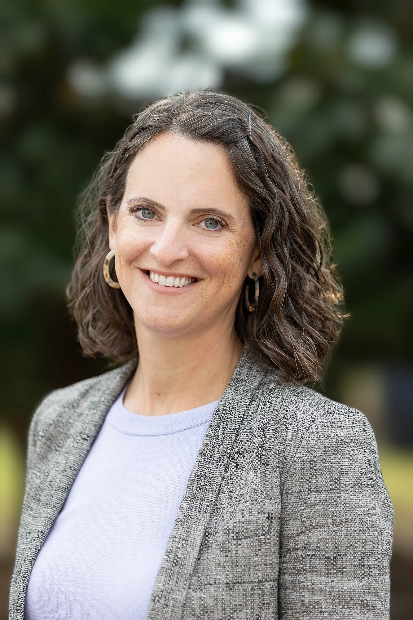 Mary Wilkinson, with shoulder-length curly brown hair, blue eyes, and wearing hoop earrings, a light gray blazer, and a white top, standing outdoors with blurred greenery in the background.