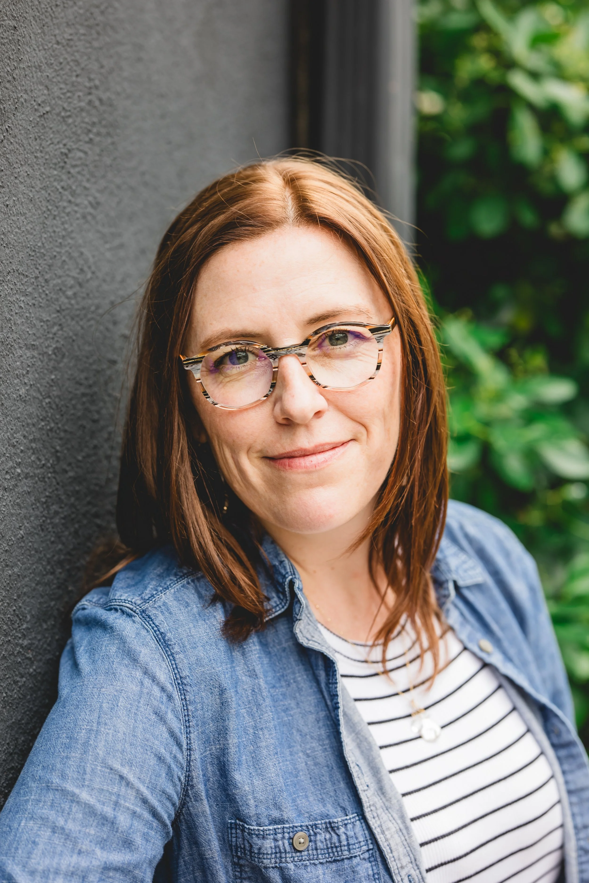 Jessica Carswell, with shoulder-length brown hair and glasses, wearing a denim jacket and a striped shirt, standing outdoors against a dark wall and greenery in the background.