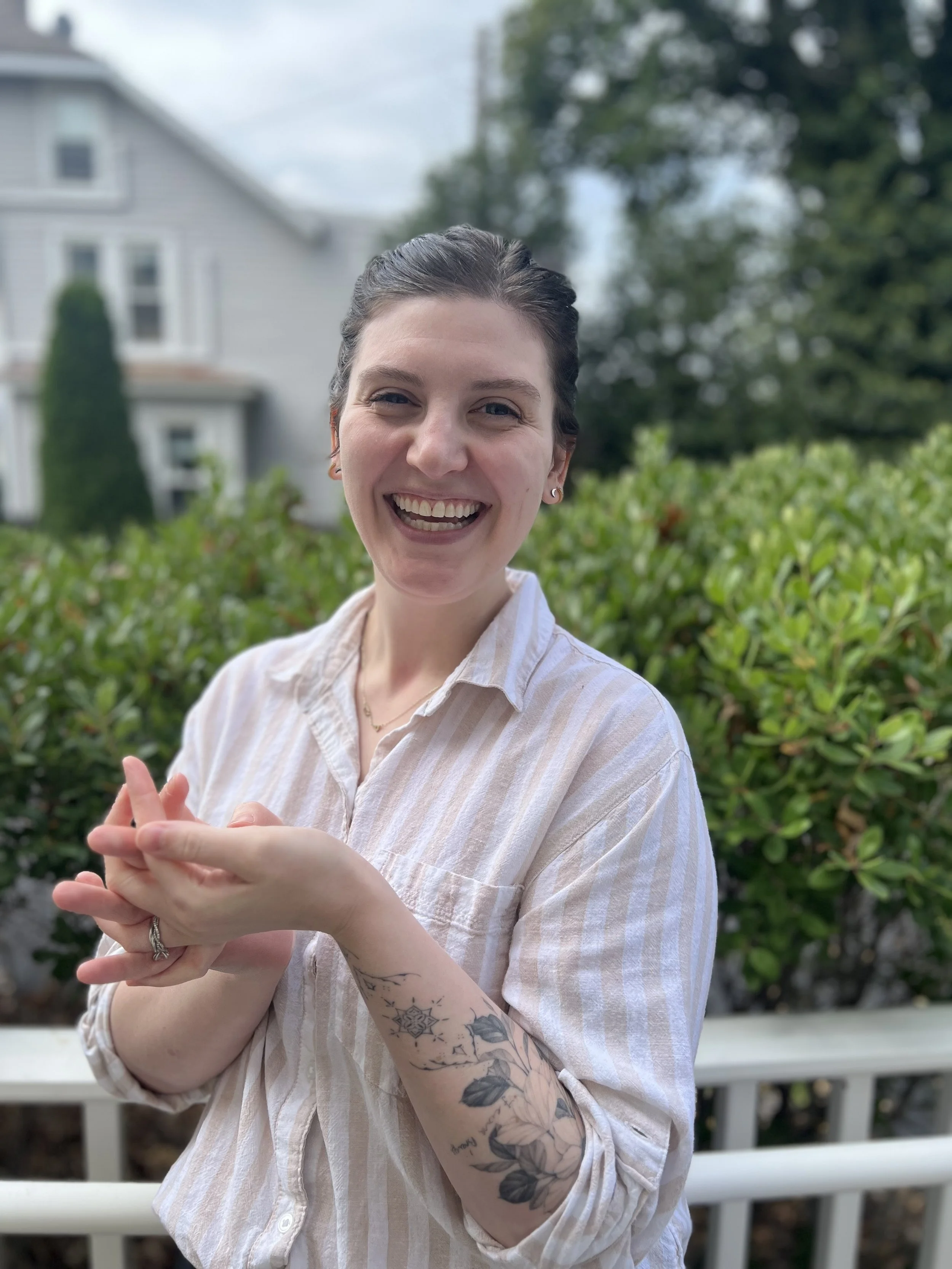 A young woman with dark hair in a bun, smiling, wearing a striped beige and white shirt, standing outdoors in front of green bushes and trees, with a house and a cloudy sky in the background.