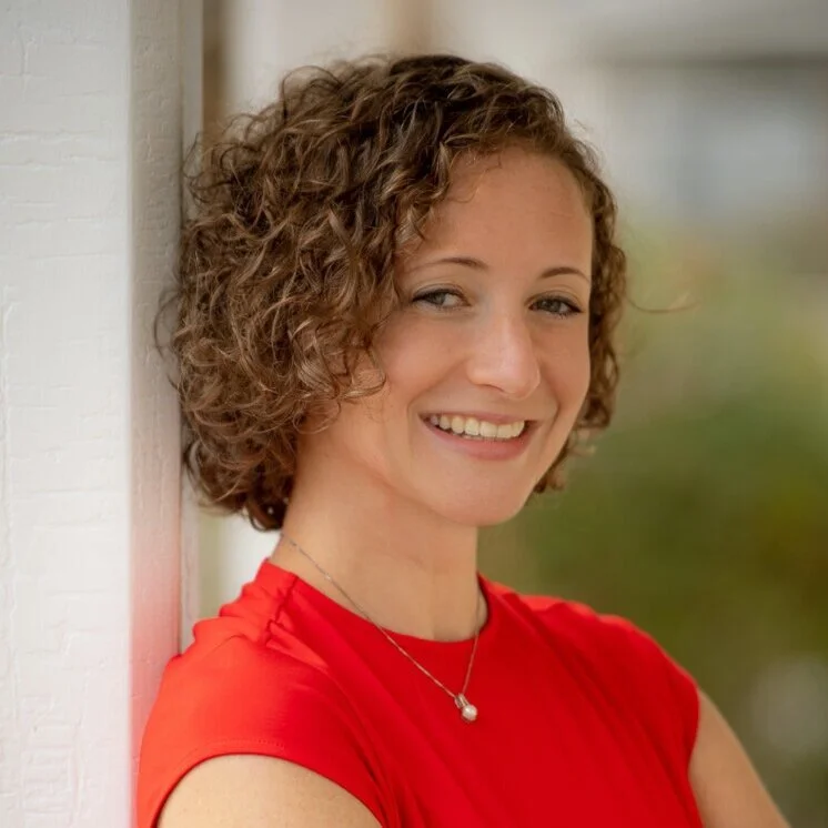 Katie Holzworth, with curly brown hair, wearing a red top and a silver necklace, smiling and leaning against a white wall outdoors.