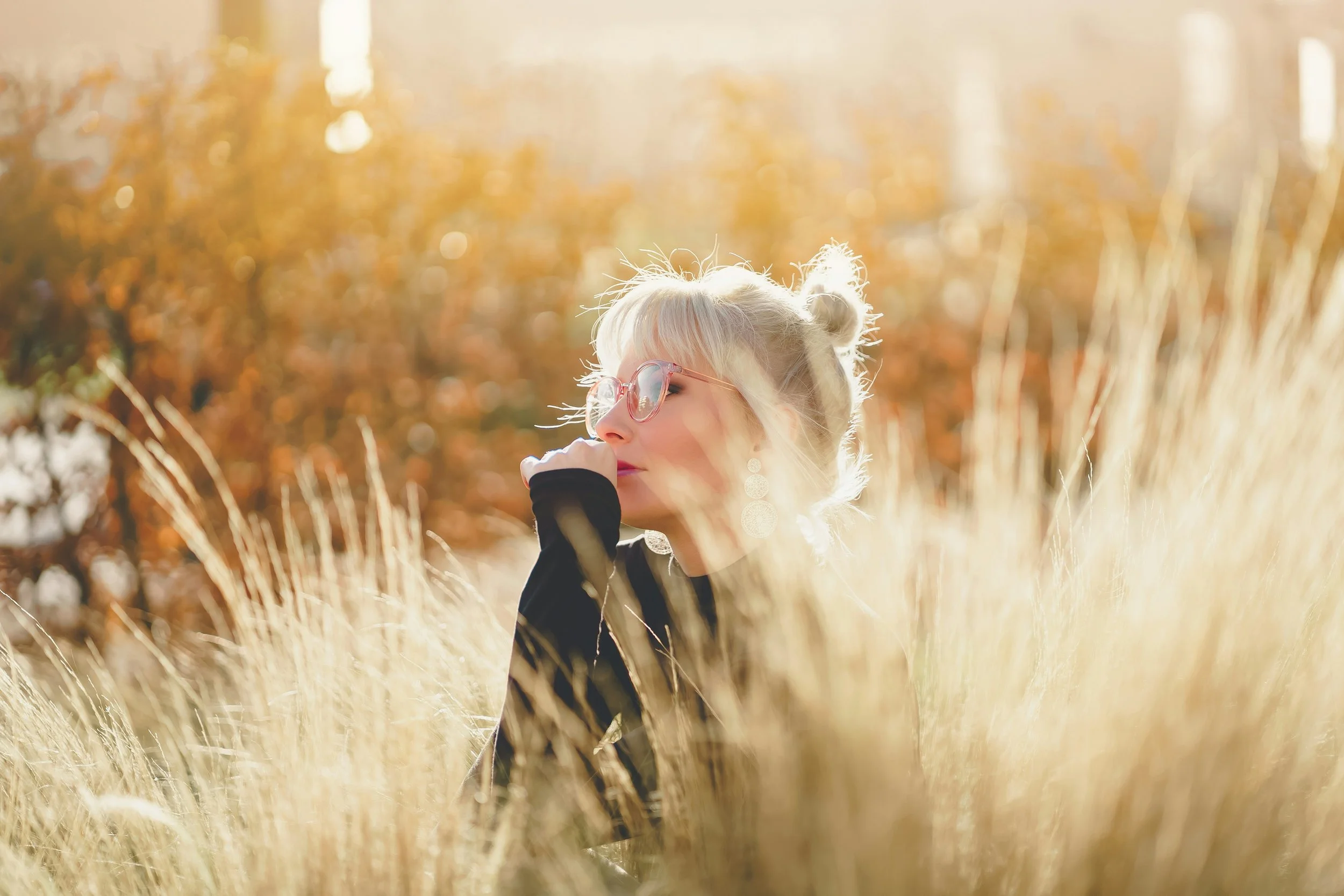 A woman with blonde hair and glasses sitting among tall grass in an autumn setting, with trees and warm sunlight in the background.