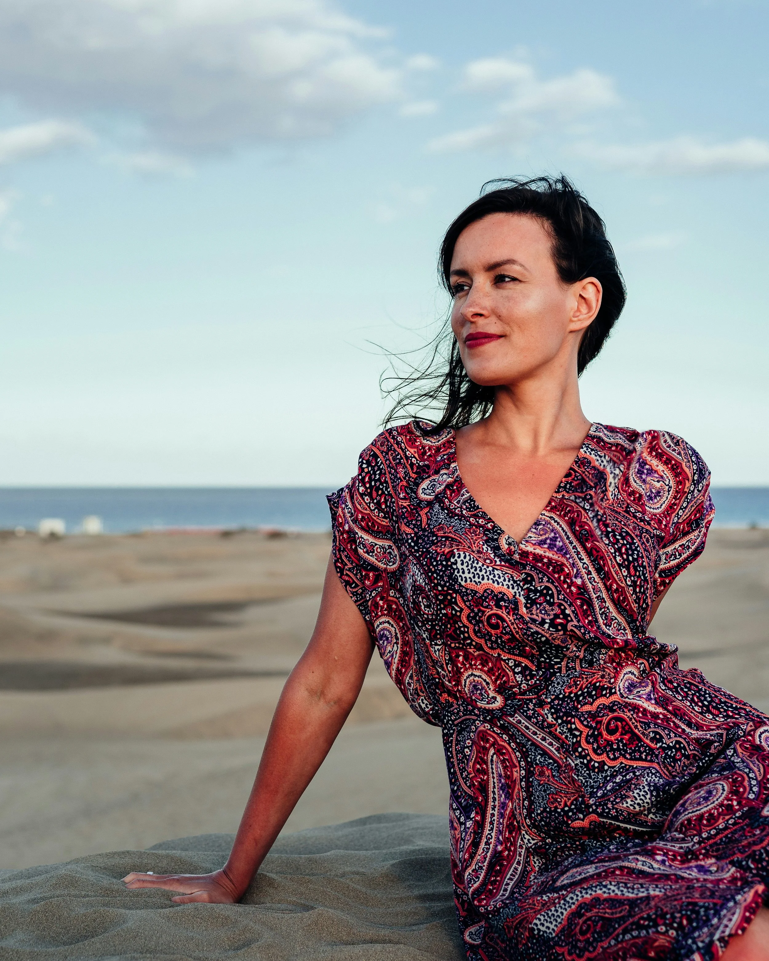Woman sitting on sand dune at beach, wearing patterned dress, with ocean and sky in background.