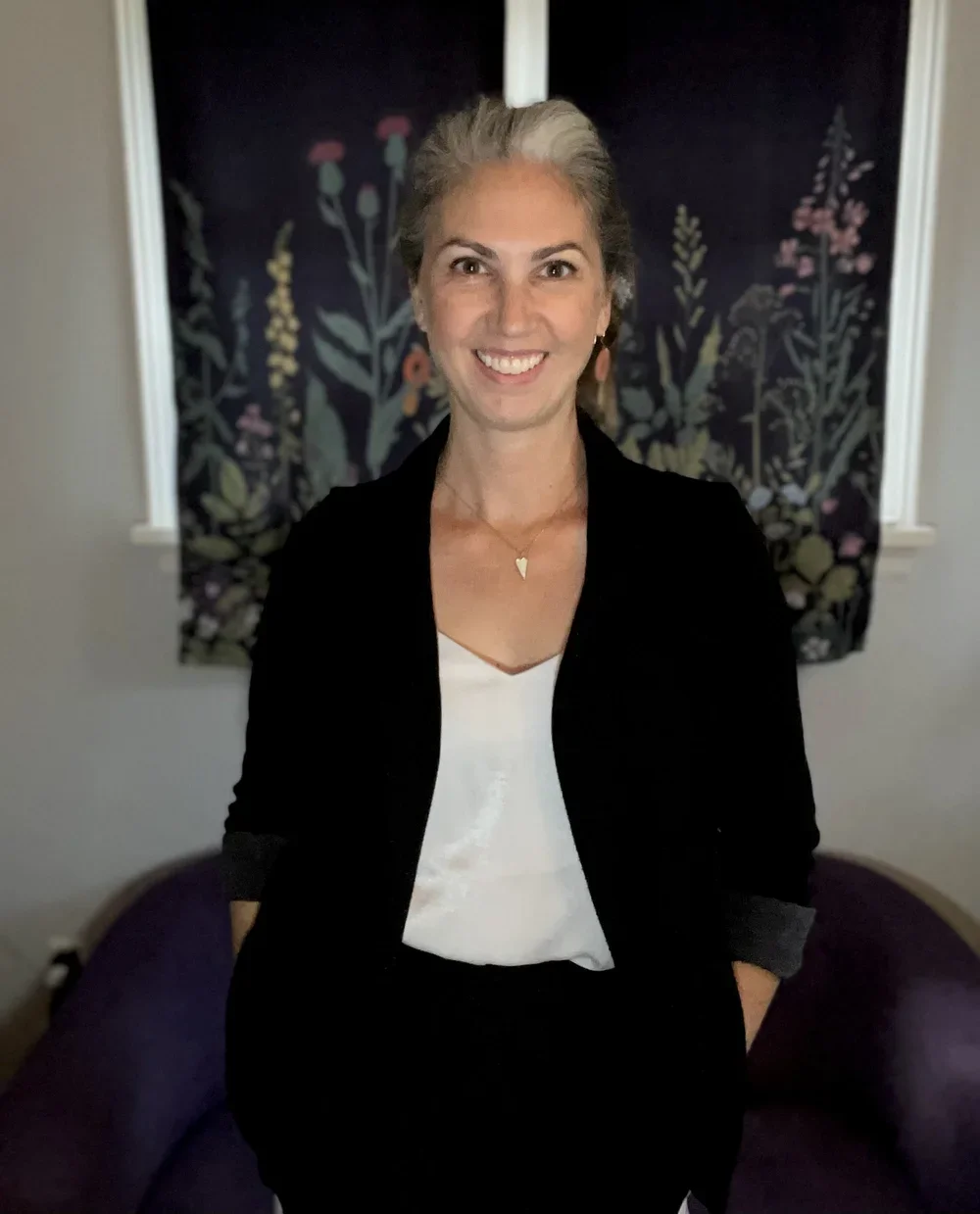 Lindsay Edwards, wearing a black blazer and white top, standing in front of a dark floral curtain.