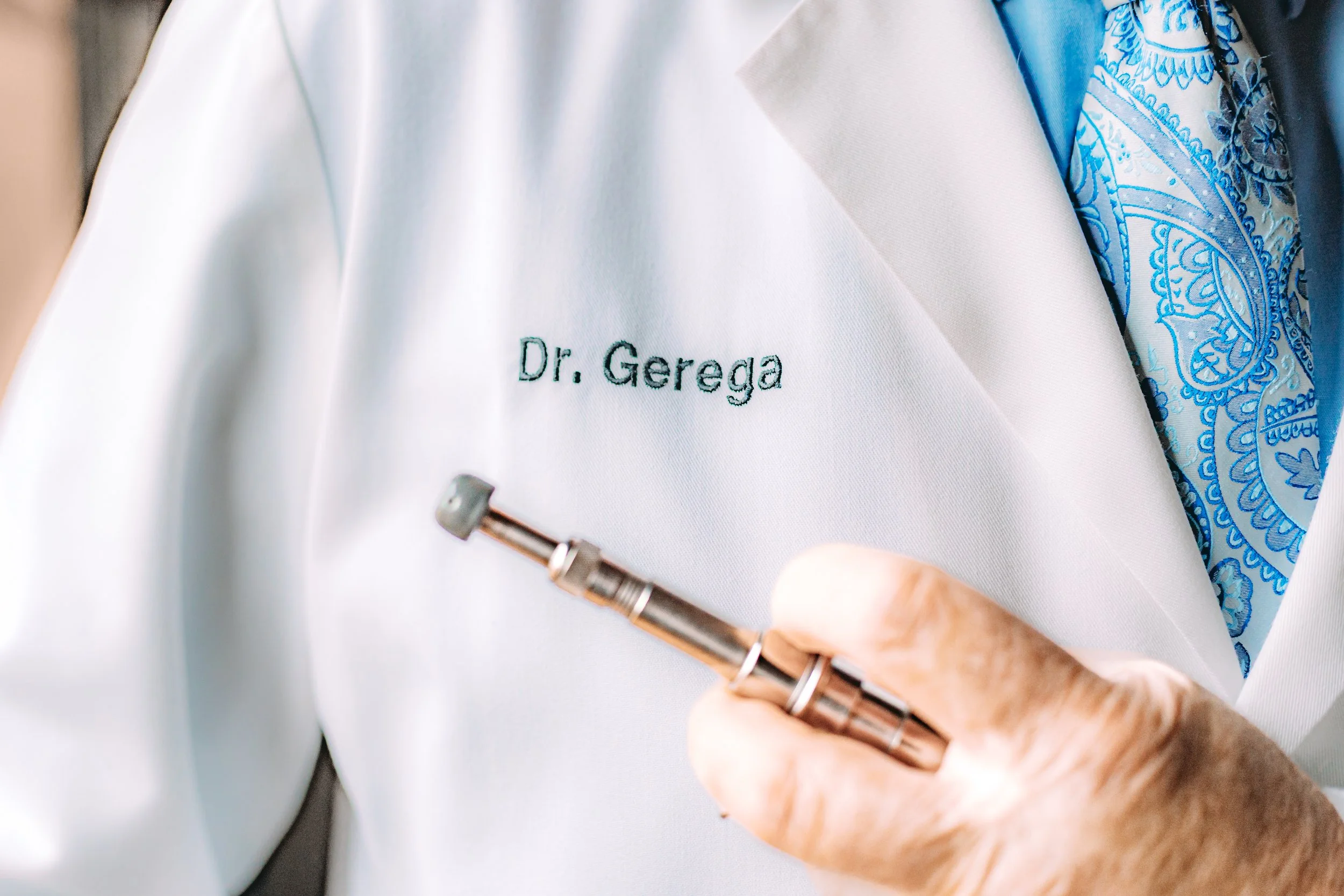 Close-up of a doctor's white coat with the embroidered name 'Dr. Gereka' and a hand holding a medical instrument, with a blue patterned scarf visible underneath.