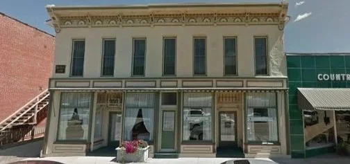 A two-story, historic building with large windows and decorative trim, situated on a street with a green storefront nearby.