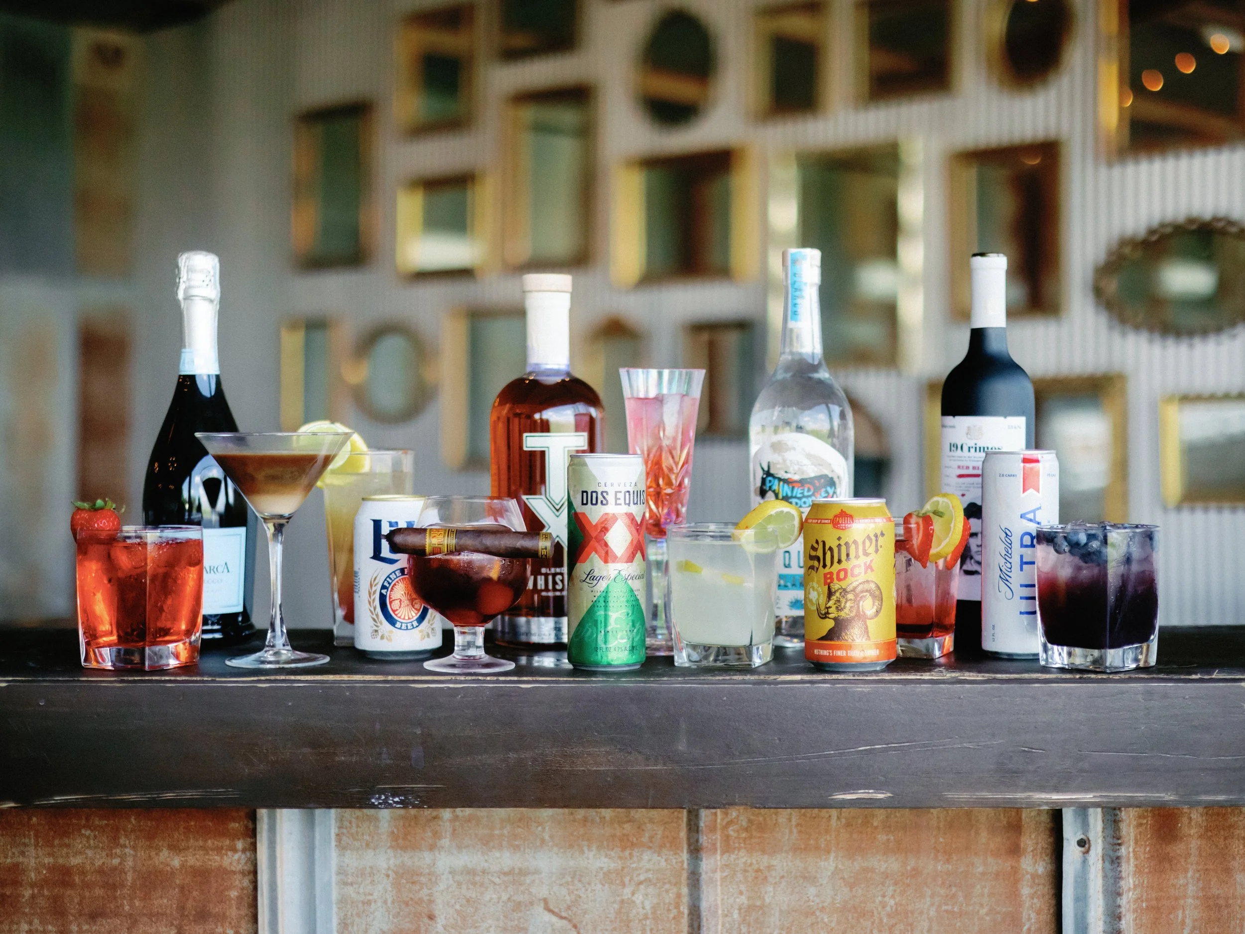 Various colorful alcoholic beverages including wine, cocktails, and cans on a dark wooden bar with bottles, some garnished with fruit slices, against a decorative wall with mirrors in the background.