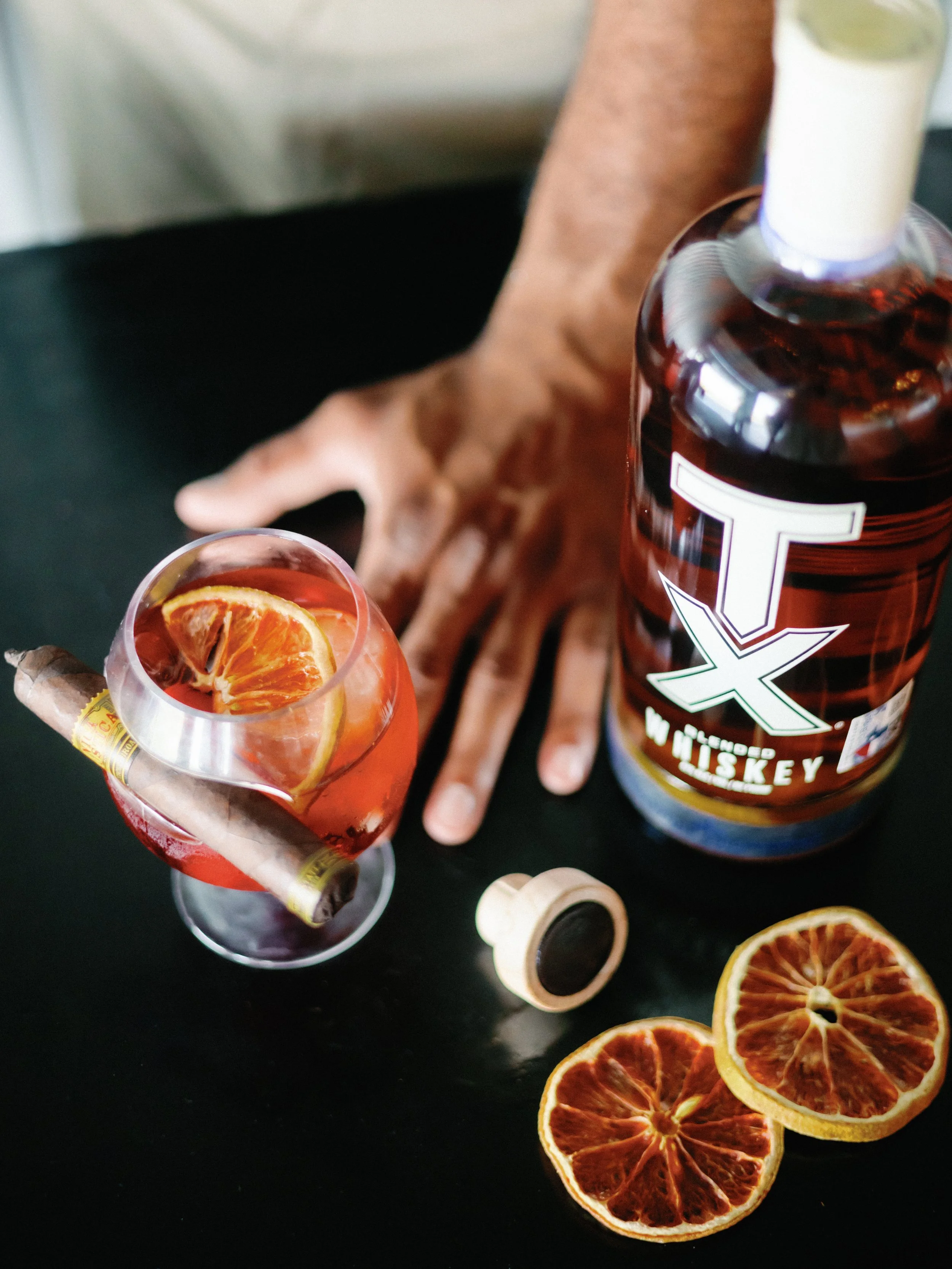 A glass of cocktail with orange slices and a cigar, a bottle of whiskey, and dried orange slices on a black table.