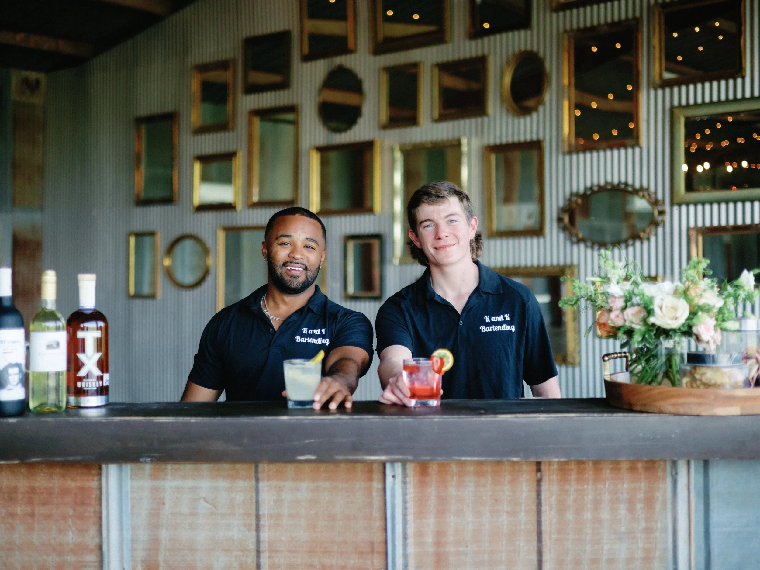 Two bartenders standing behind a bar counter with drinks, bottles, and flowers, smiling at the camera against a backdrop of wall mirrors.