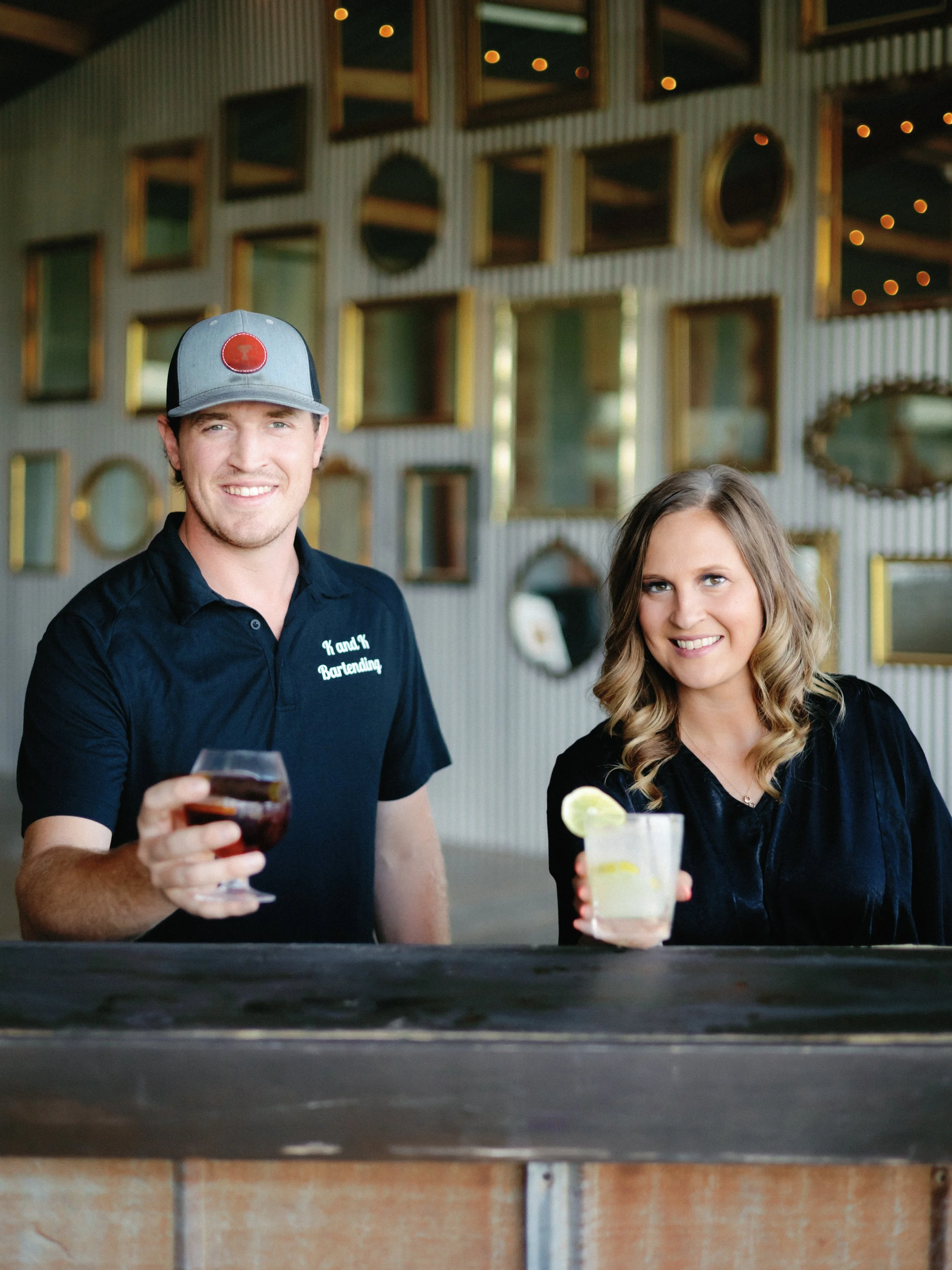 A man and woman sitting at a bar holding drinks, smiling at the camera, with a decorative mirror wall in the background.