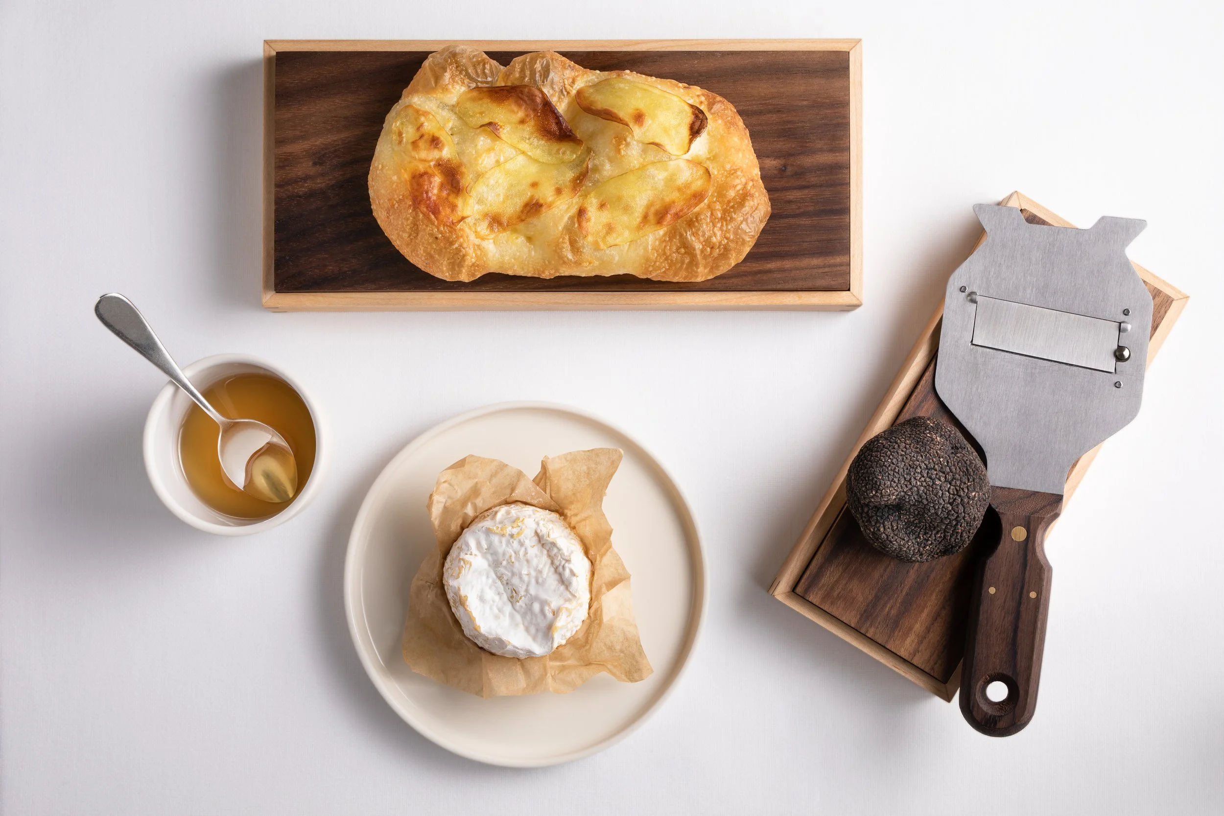 A table with a loaf of bread, a soft cheese on a cream-colored plate, honey in a white cup with a spoon, and a black truffle on a wooden-handled cheese slicer.