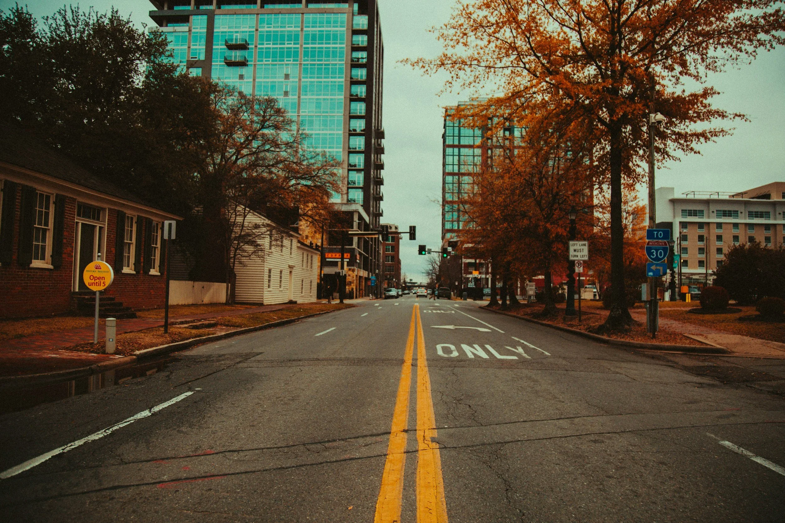 City street with tall modern buildings, trees with orange fall leaves, and traffic lights, shot from the perspective of a car at an intersection with yellow double lines, and painted road markings that say 'ONLY' for a turn or lane restriction.