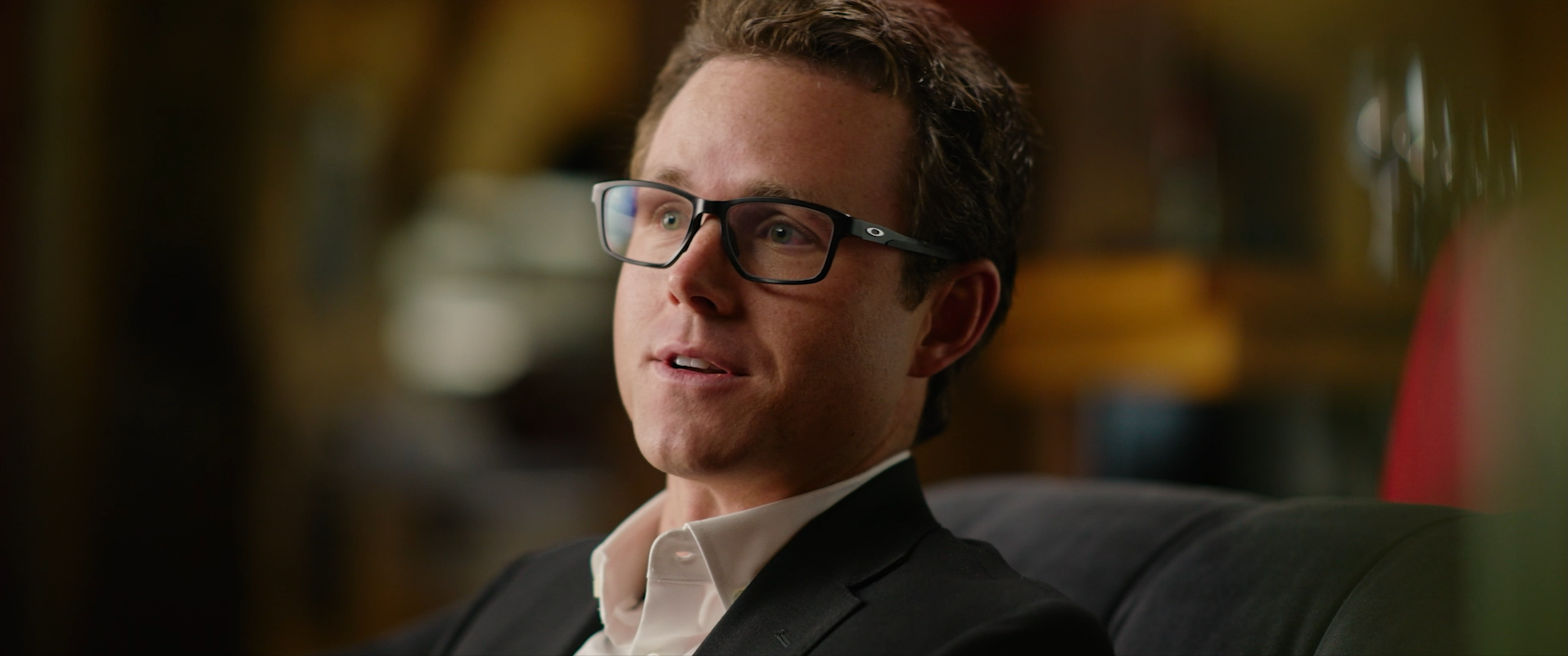 A man wearing glasses and a black suit sitting on a chair in an indoor setting with warm lighting.