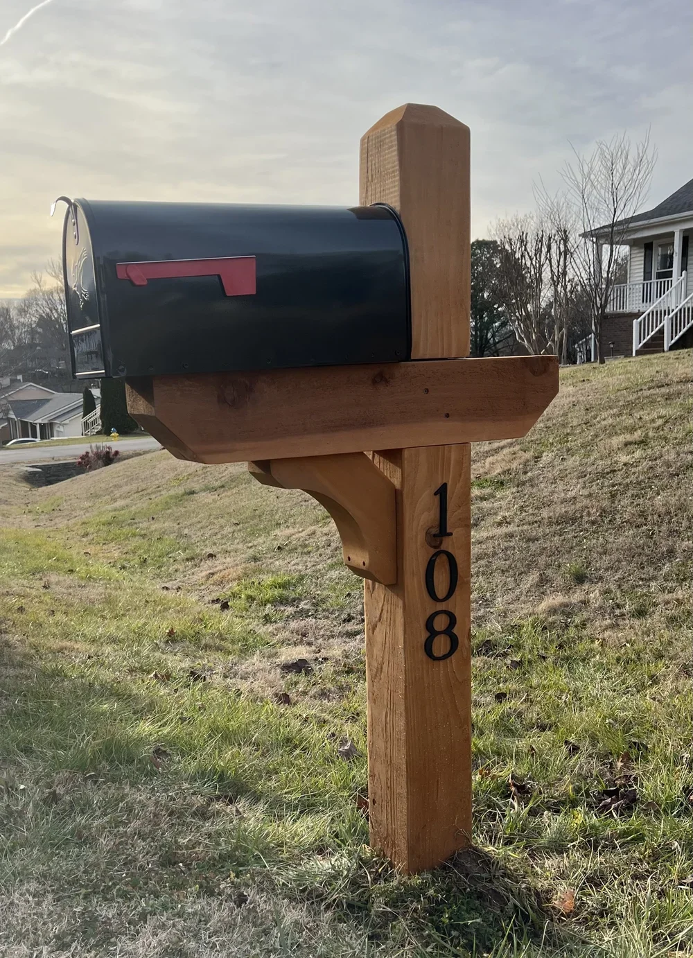 A black mailbox with a red flag mounted on a wooden post with the house number 108, situated on a grassy lawn in a suburban neighborhood.