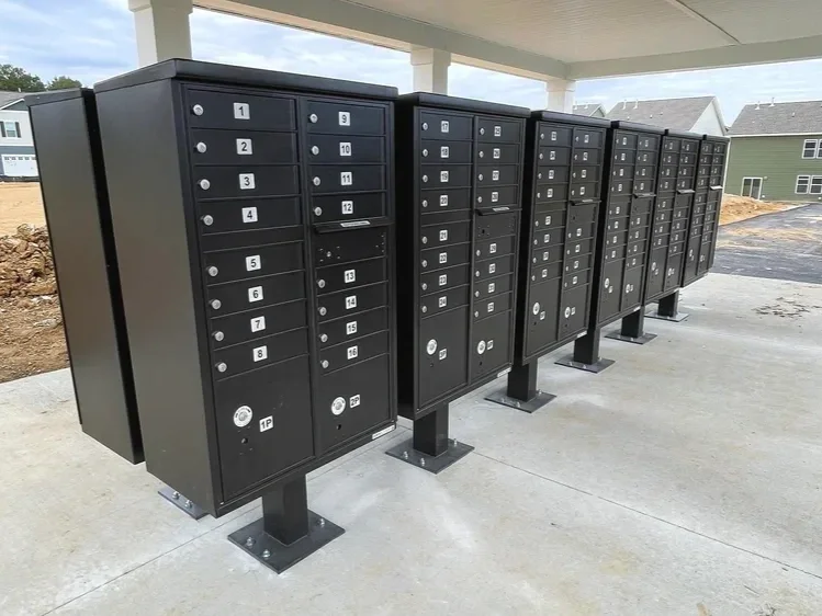 A row of black individual mailboxes with numbered compartments, located outdoors on a concrete patio under a roofed area, with houses and ground under construction in the background.
