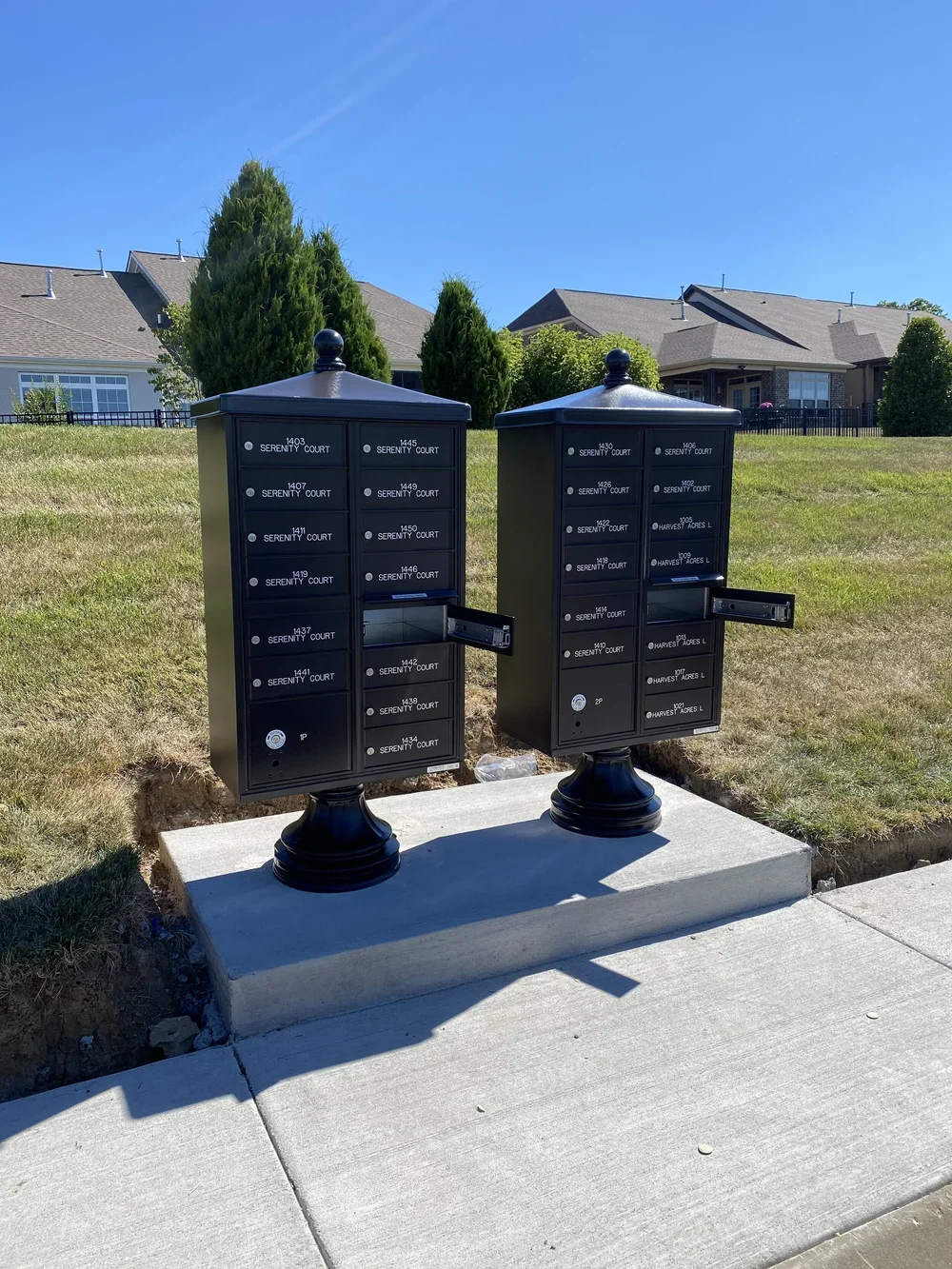 Two black parcel mailboxes labeled Serenity Court and Harvest Acres L, mounted on concrete base on sidewalk in residential neighborhood, with houses and trees in background.