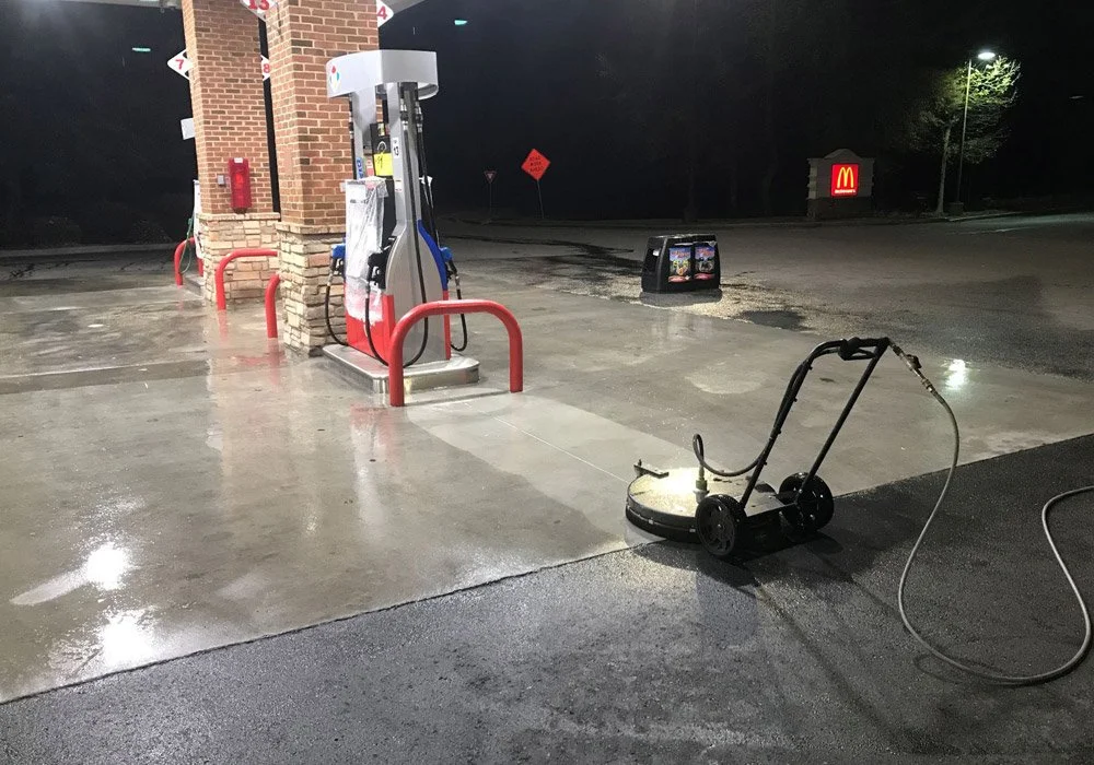 A gas station at night with a power scrubber cleaning the concrete pavement, with a McDonald's sign visible in the background.