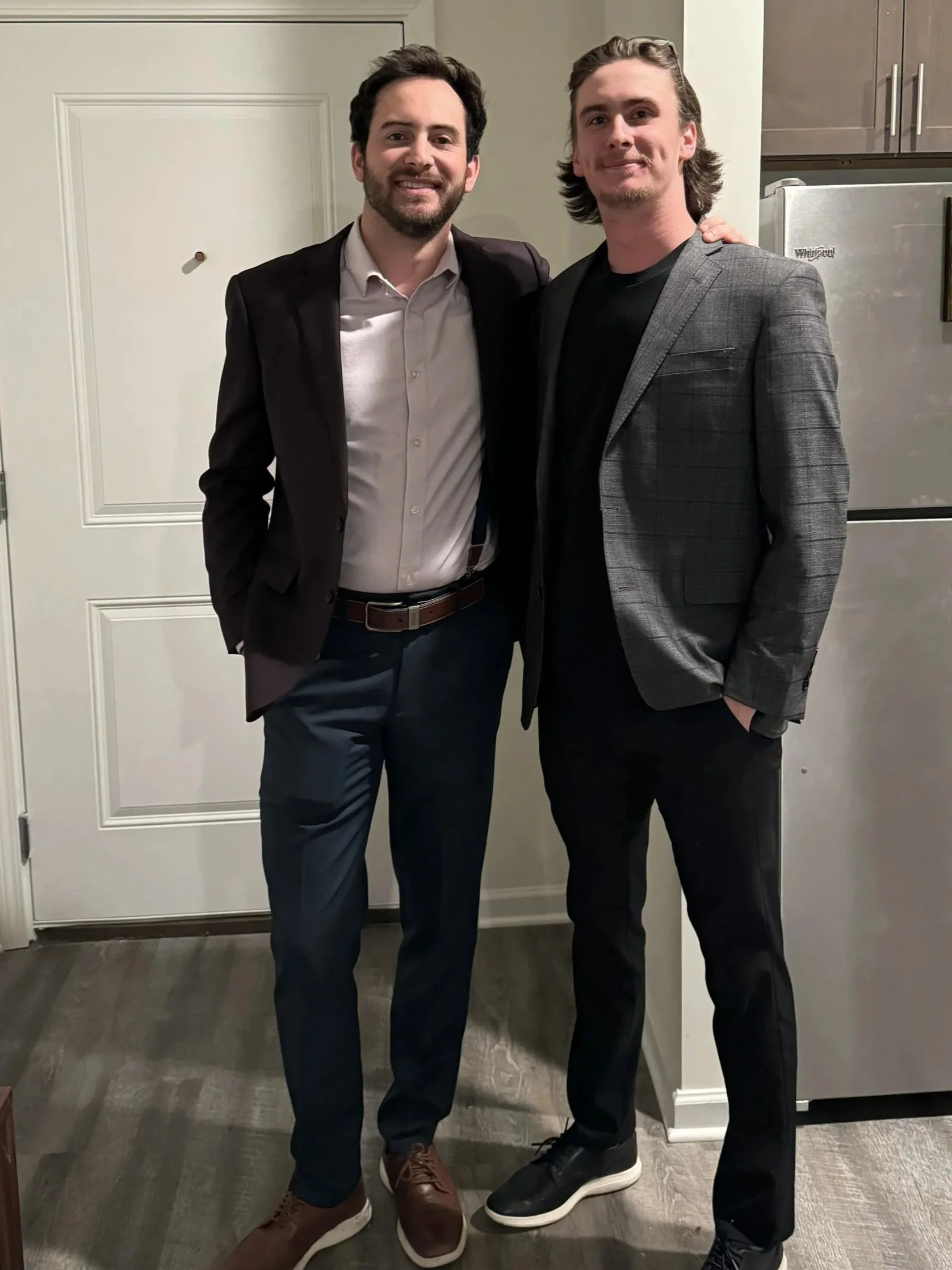 Two men dressed in suits standing together indoors, smiling with one man's arm around the other. One man has dark hair and a beard, wearing a black blazer over a white shirt. The other has long blonde hair, wearing a gray checked blazer over a black shirt.