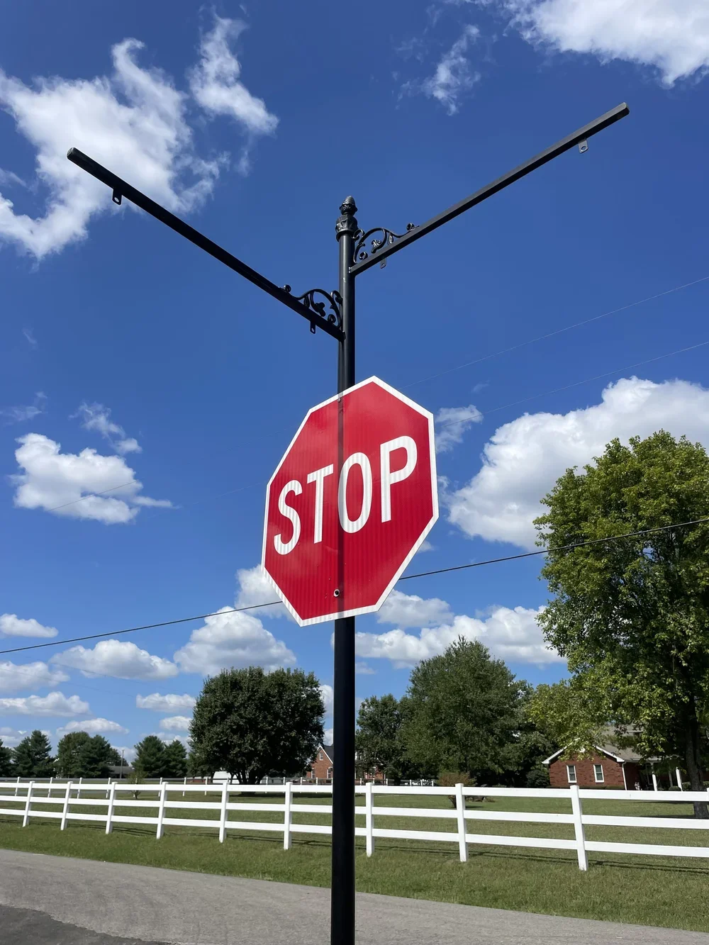 A stop sign attached to a decorative black pole with an ornate arm, set against a bright blue sky with white clouds. Green trees and a white fence are in the background.