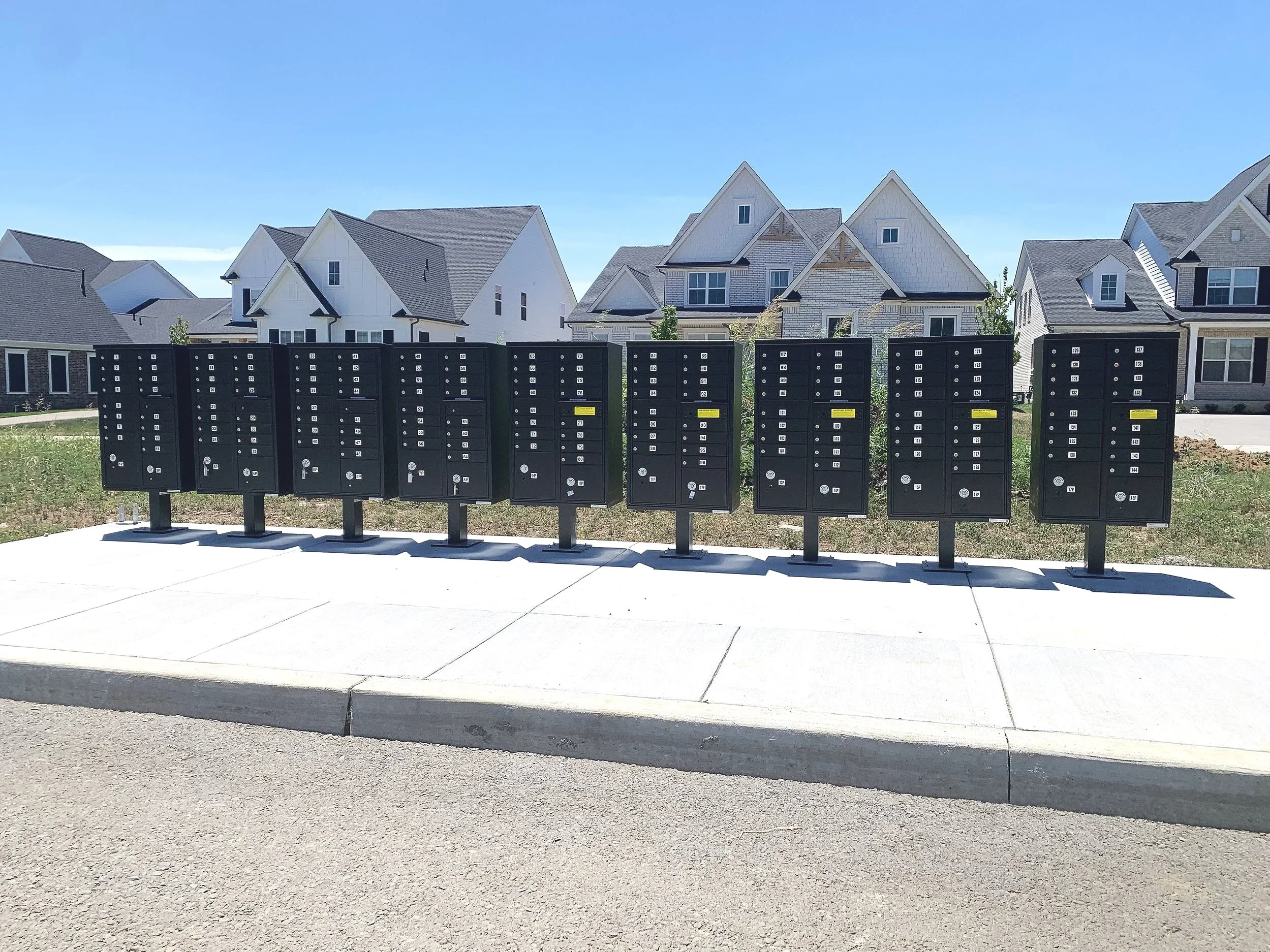 Multiple black mailbox units with white labels in a row in a residential neighborhood with new houses in the background under a clear blue sky.