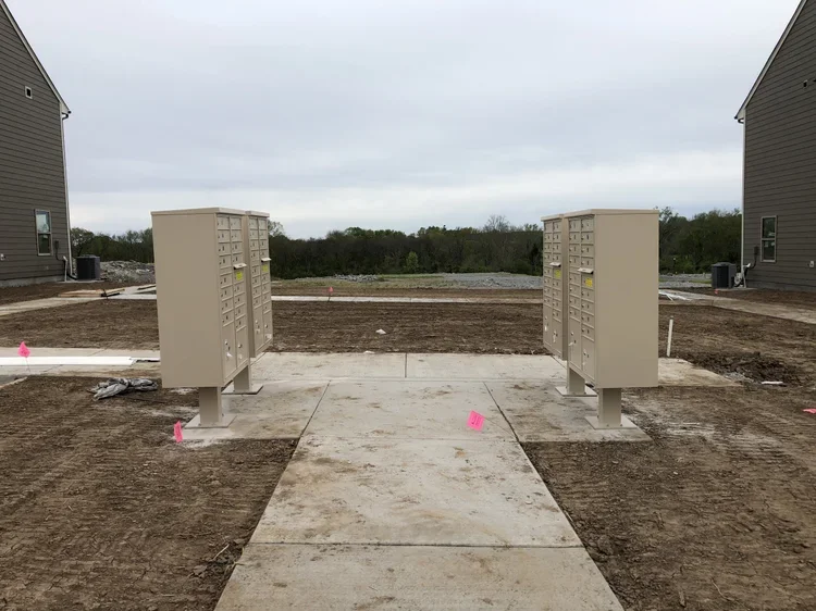 Two beige apartment mailboxes installed on concrete slabs in an outdoor construction site with dirt and gravel ground, flanked by residential buildings and surrounded by an open field under a cloudy sky.
