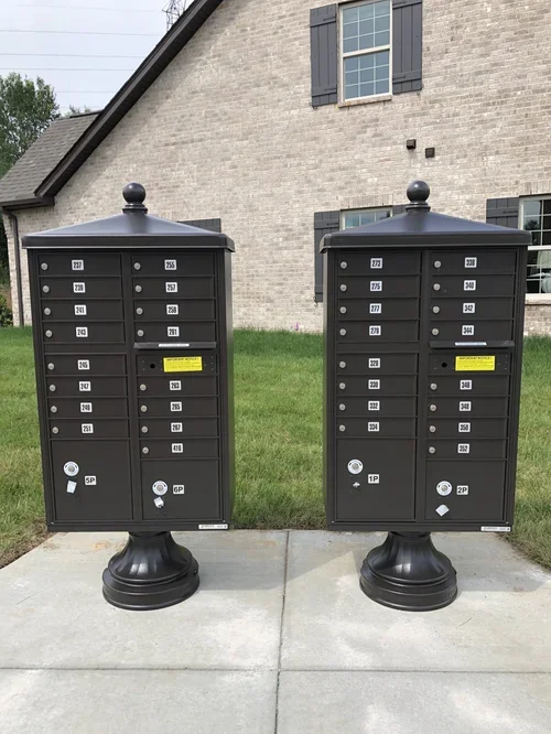 Two black outdoor mailboxes with multiple compartments, labeled with apartment numbers, standing on a concrete sidewalk in front of a house with brick walls and windows.