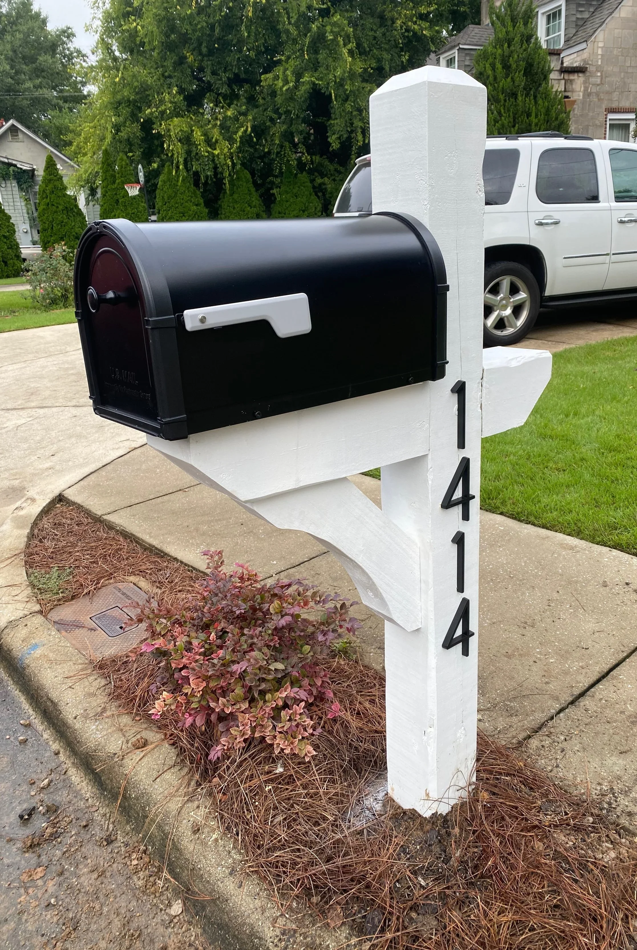 A black mailbox mounted on a white wooden post with house number 1414, next to a small plant and a sidewalk.