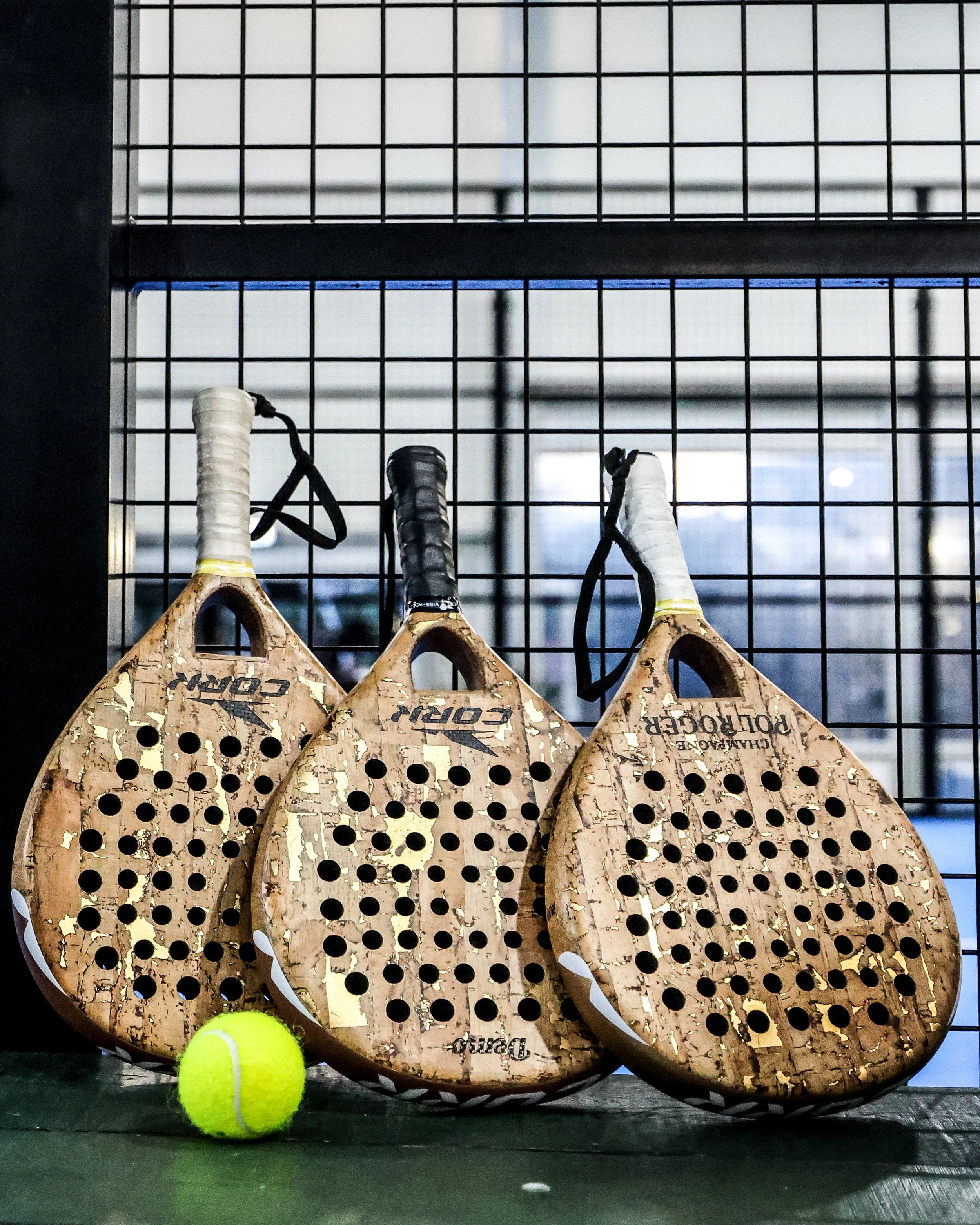 Three cork pickleball paddles with black and white grips leaning against a black wire fence, with a yellow tennis ball in front of them. 