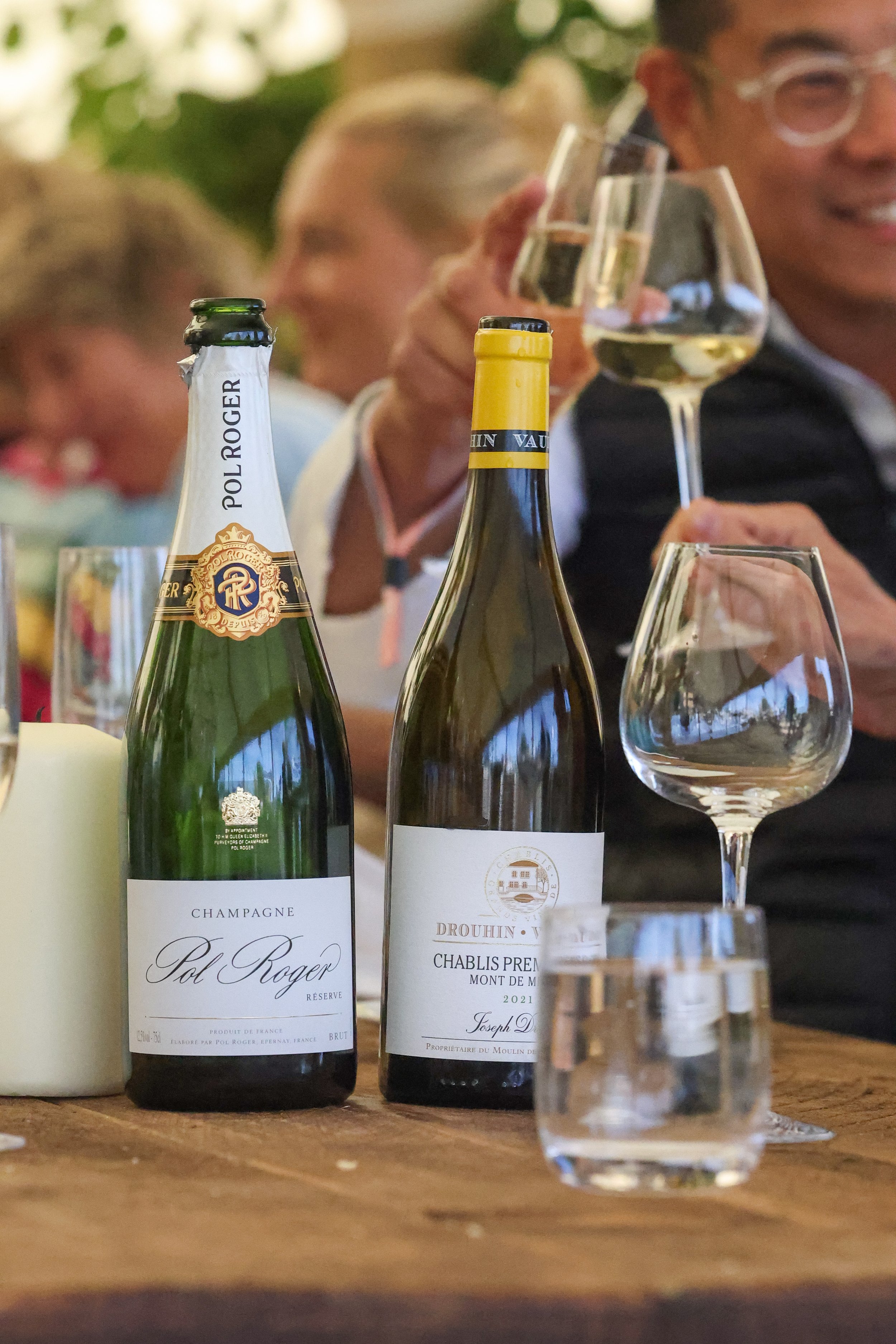 Two bottles of Pol Roger champagne and a bottle of white wine on a wooden table, with glasses and people celebrating in the background.