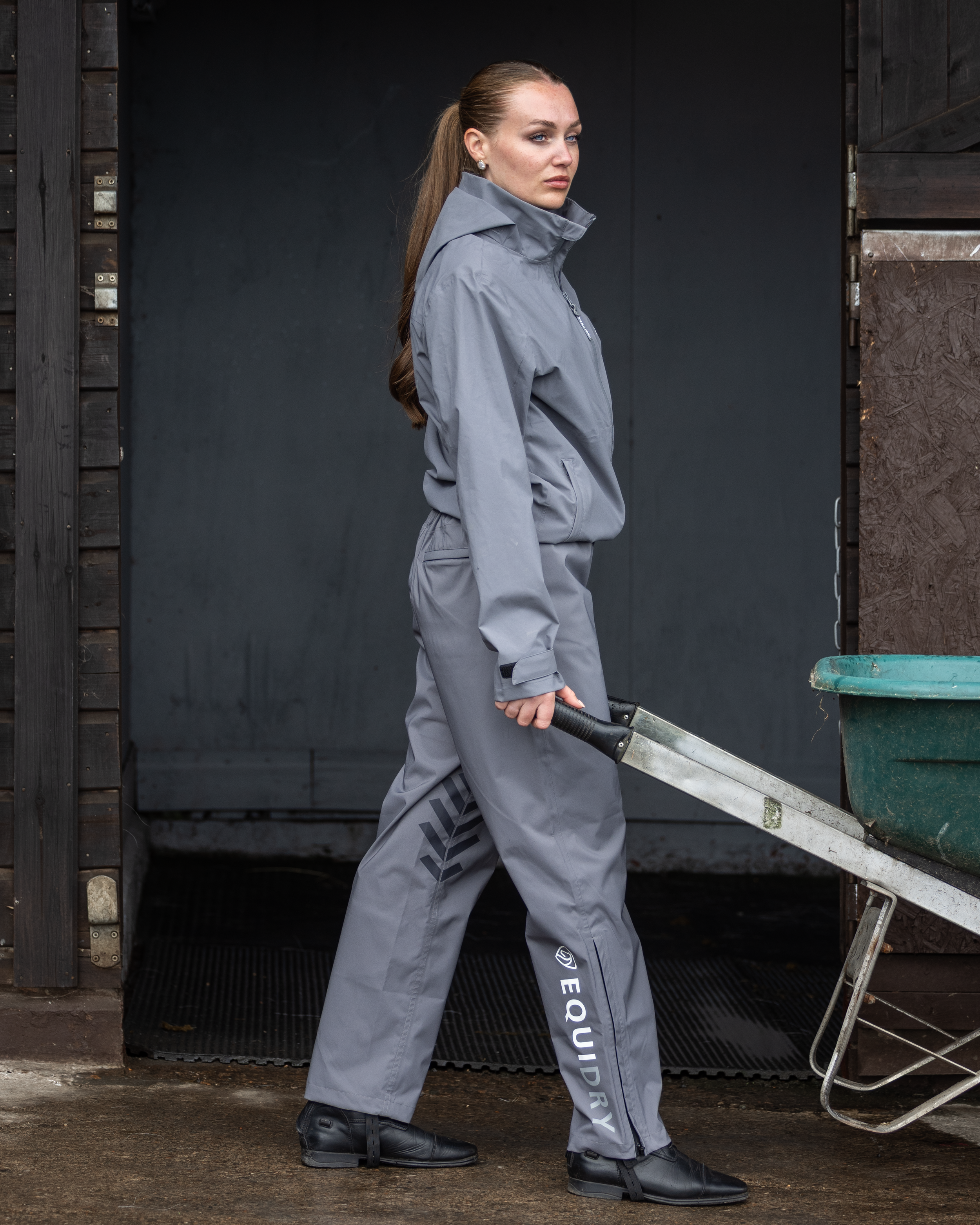 Woman in grey waterproof trousers pushing a wheelbarrow outside a horse stable.