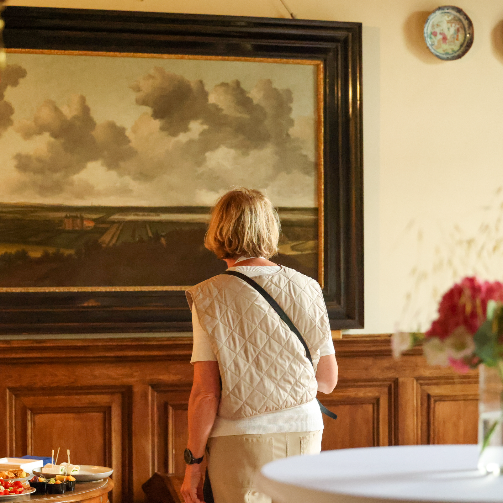 A woman with short blonde hair wearing a white quilted vest and cream-colored pants is looking at a large landscape painting in a room with wooden paneling. Part of a table with a flower arrangement and some food is visible in the foreground.