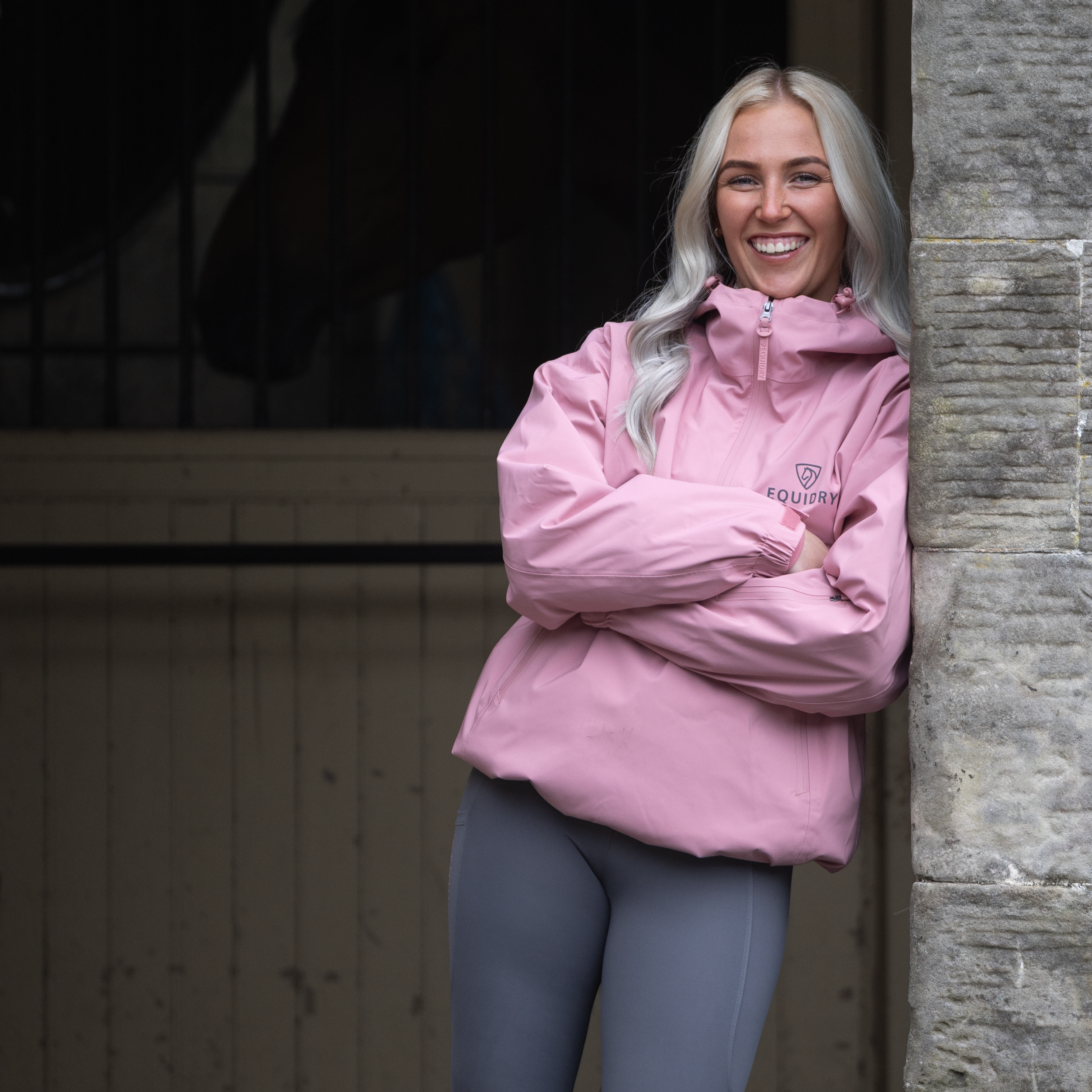 A smiling woman with long, light-colored hair leaning against a stone wall, wearing a pink jacket and gray leggings.