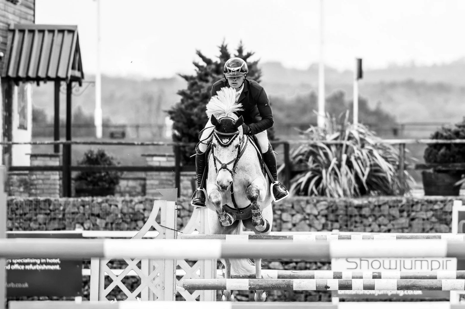 A male equestrian athlete in formal riding attire, including a helmet, riding a horse with a white mane, jumping over an obstacle during a show jumping competition at the David Broome Event Centre.