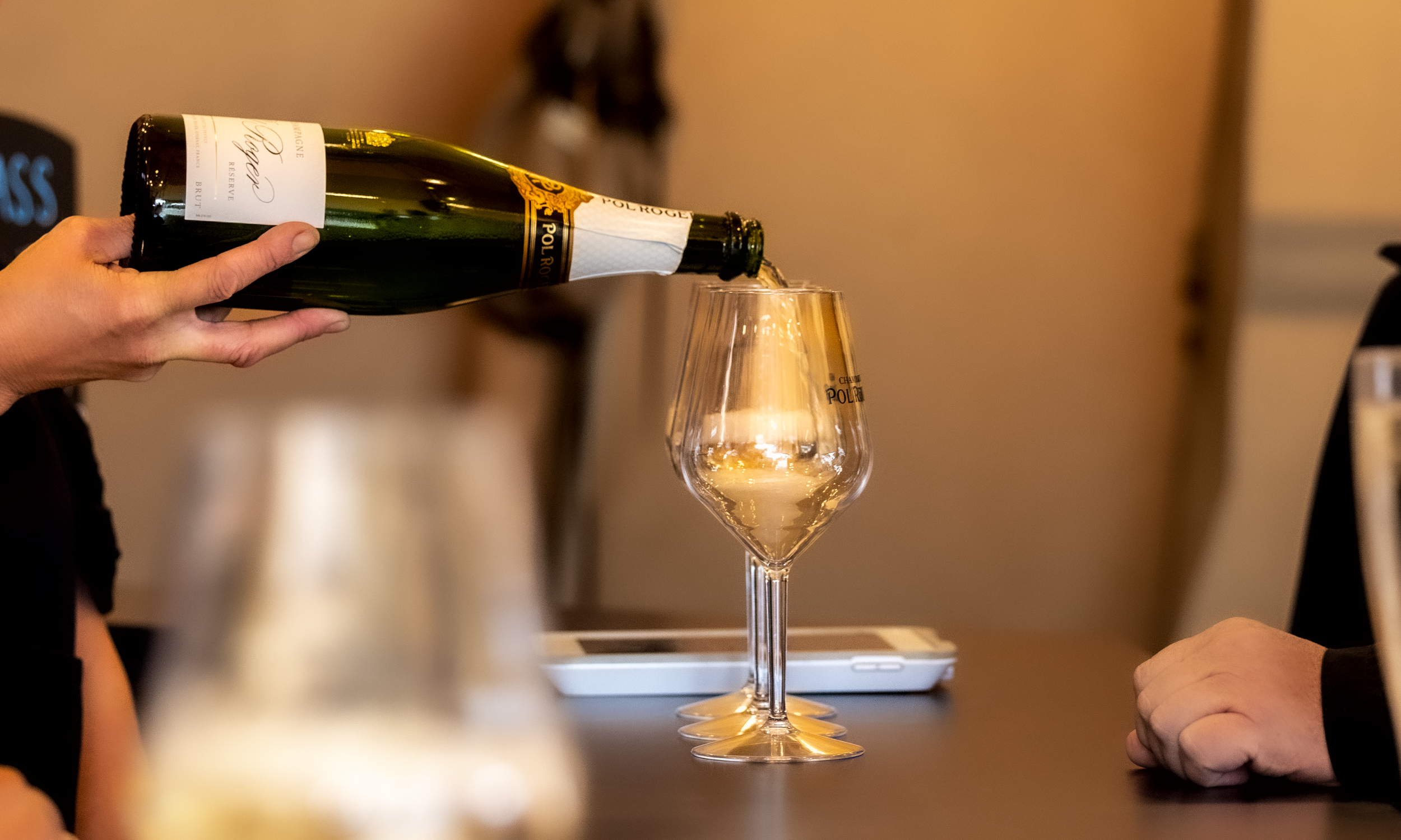 Person pouring Pol Roger champagne into a glass, with others sitting at a table, and a blurry bottle in the foreground.