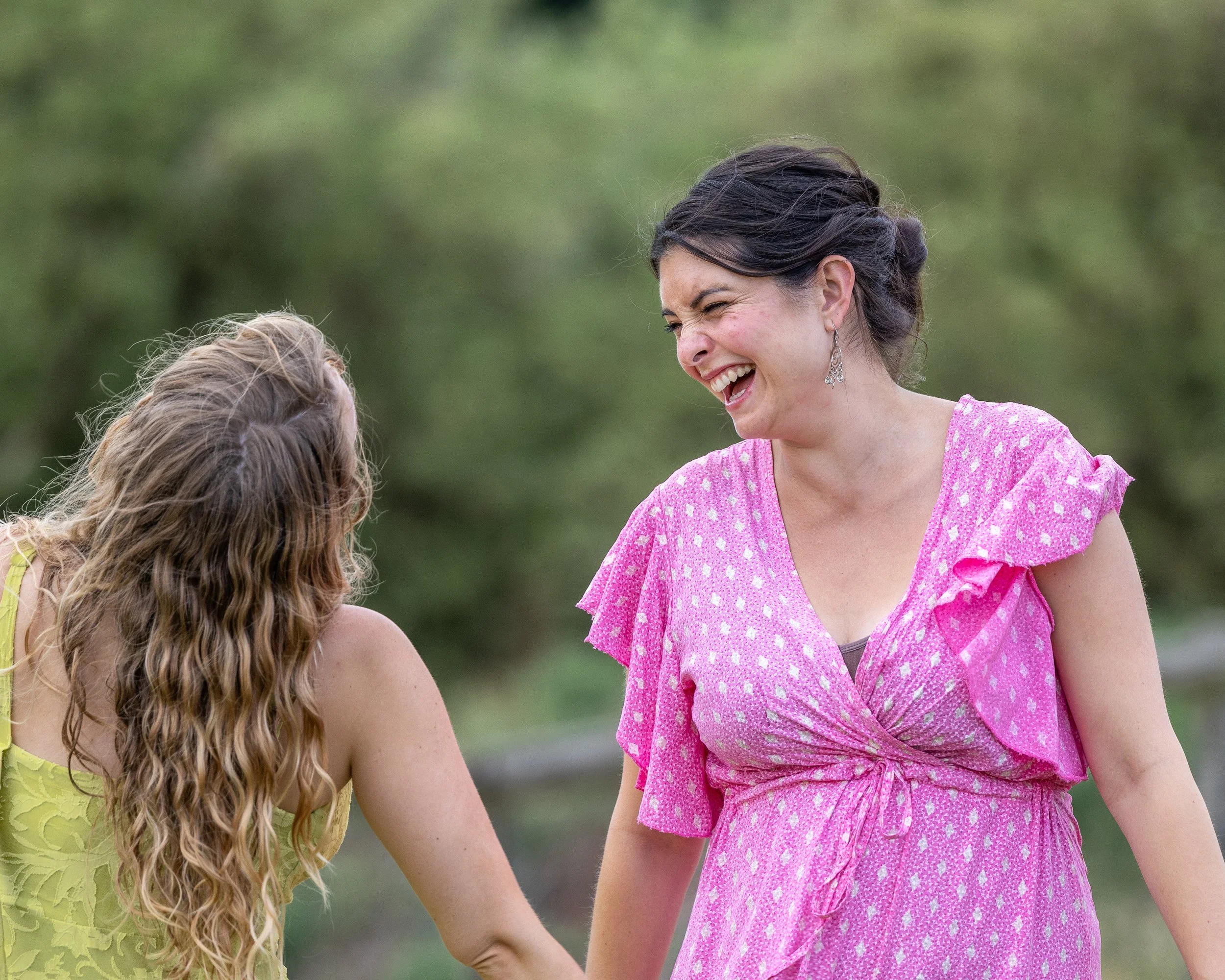 Two women holding hands and laughing outdoors with green trees in the background.