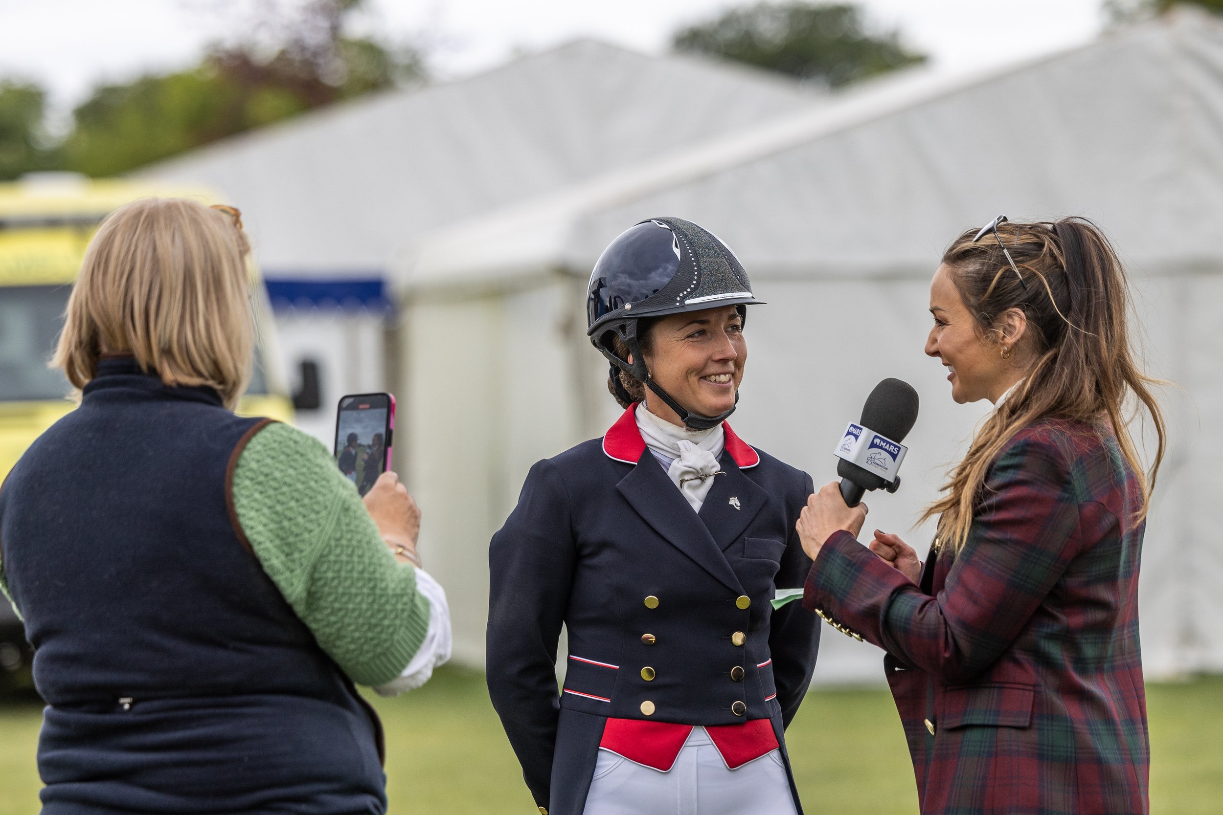 Three women are outdoors, one is interviewing another in riding attire; the woman being interviewed is smiling and wearing a helmet, while the interviewer holds a microphone, and the third woman is taking a photo with her phone.