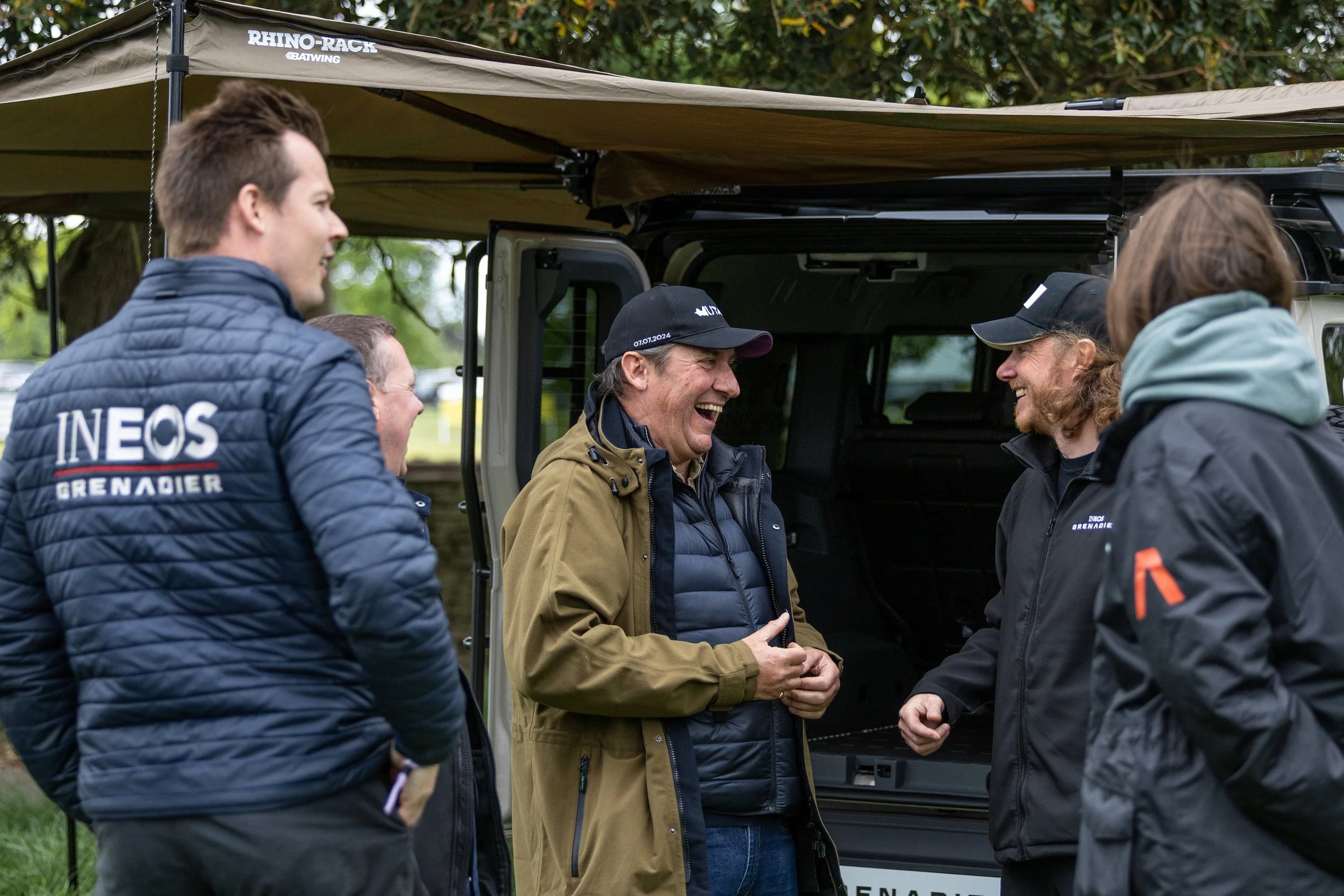 Group of five people standing outdoors, smiling and engaged in conversation, near a vehicle with an open back, under a canopy tent labeled 'Rhino-Rack'. The people are dressed in outdoor jackets, some with logos reading 'INEOS Grenadier'.